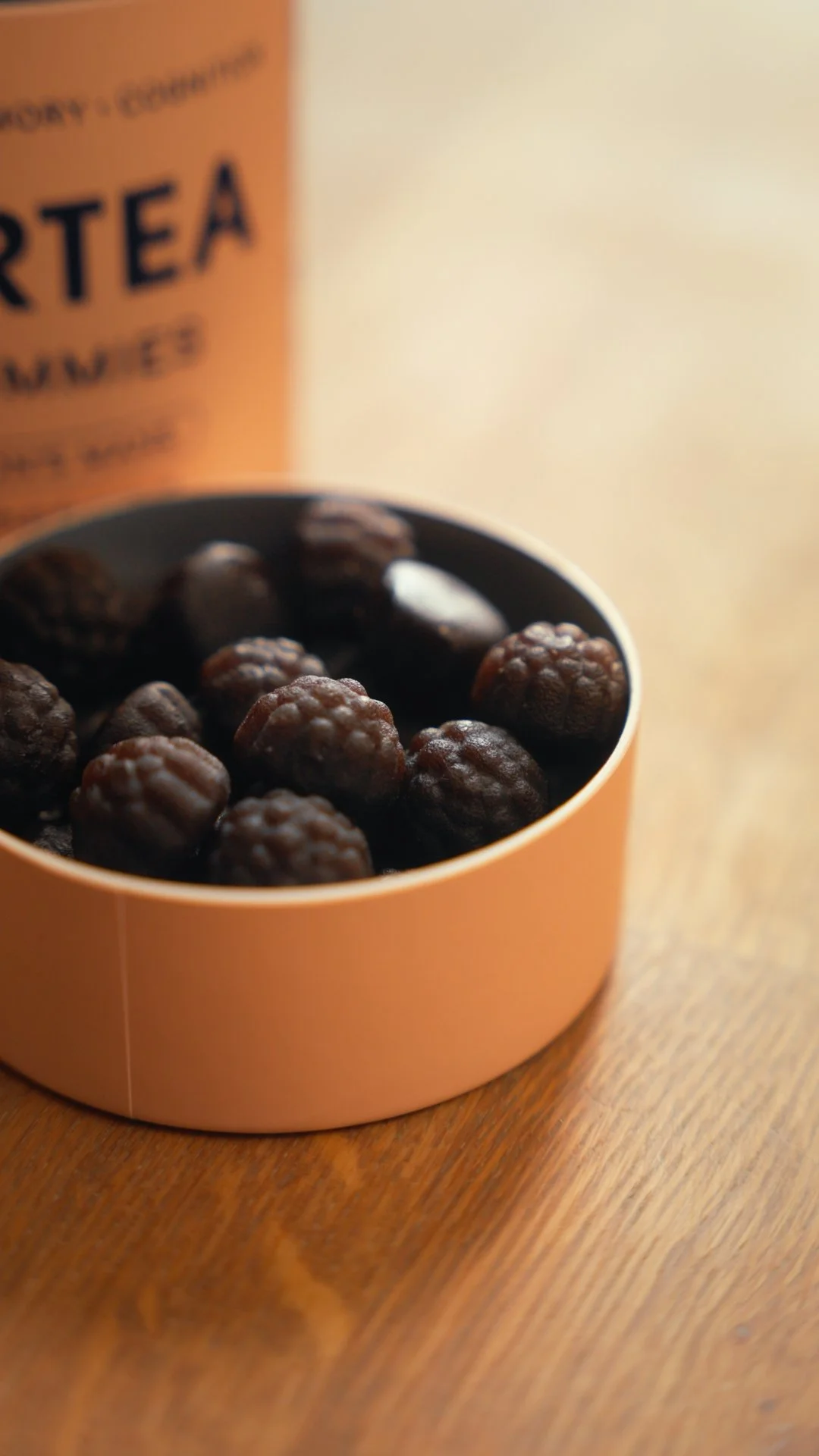Close-up of a pink container filled with blackberries on a wooden surface, with a blurred orange container labeled "TEA" in the background.