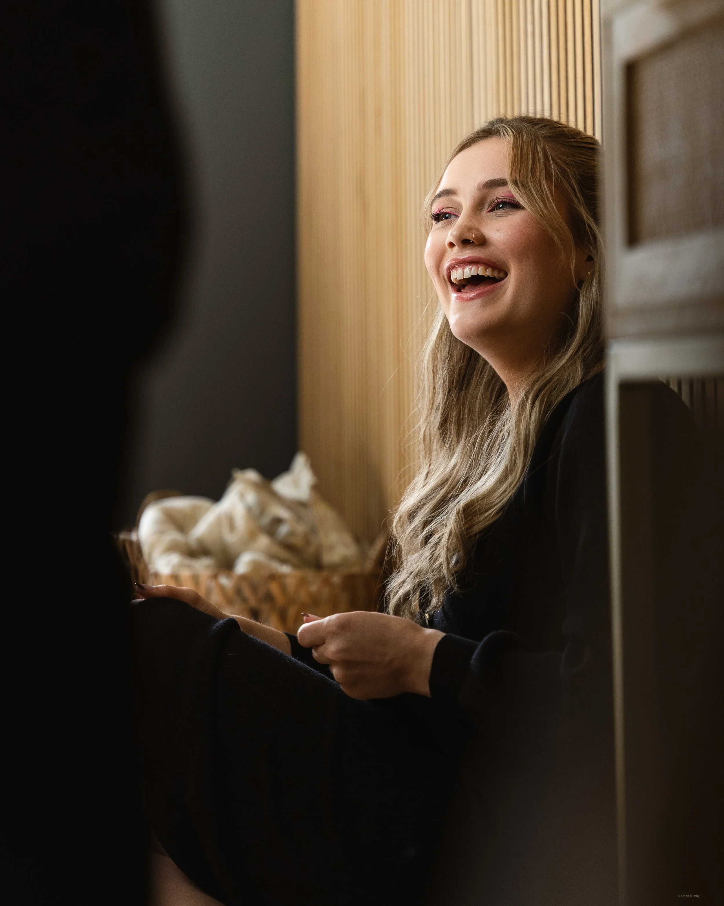 A smiling young woman with long hair, makeup, and a nose piercing, sitting against a wooden panel wall.