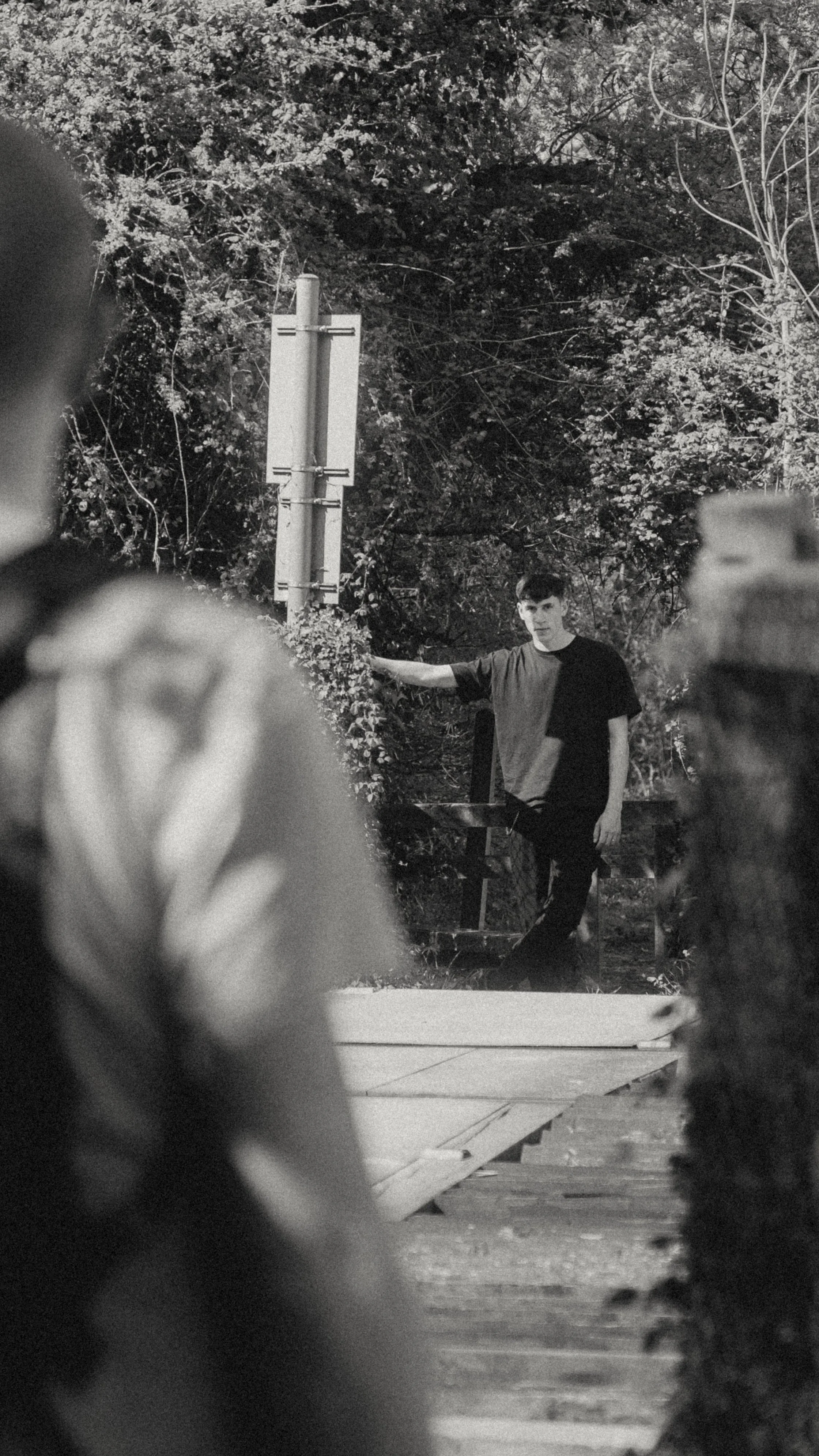 Black and white photograph of a young man standing outdoors among trees and foliage, leaning with one arm on a signpost, wearing a t-shirt, with a focused expression.