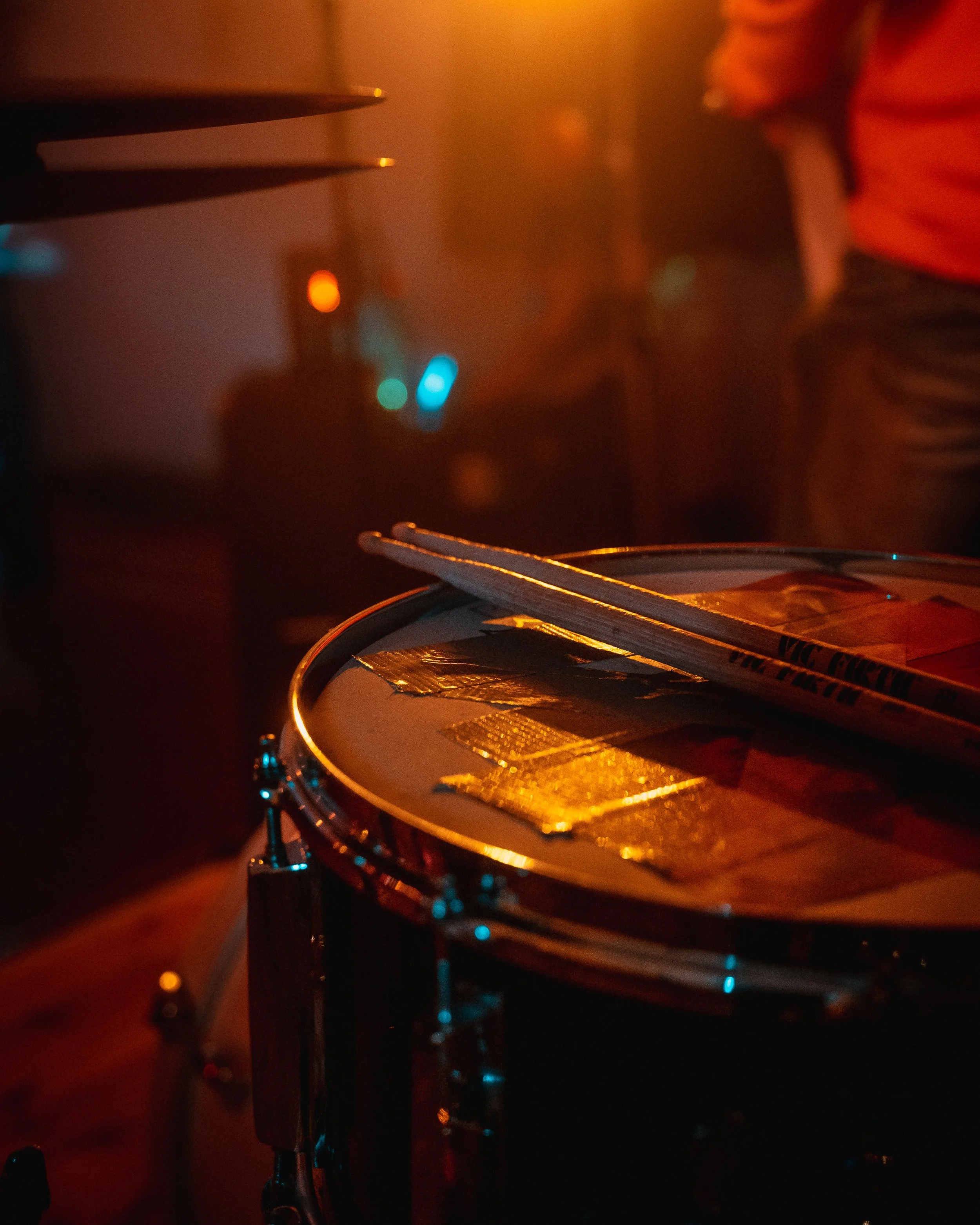 Close-up of a snare drum with drumsticks resting on top, in a dimly lit, warm-colored setting with a person partially visible in the background.