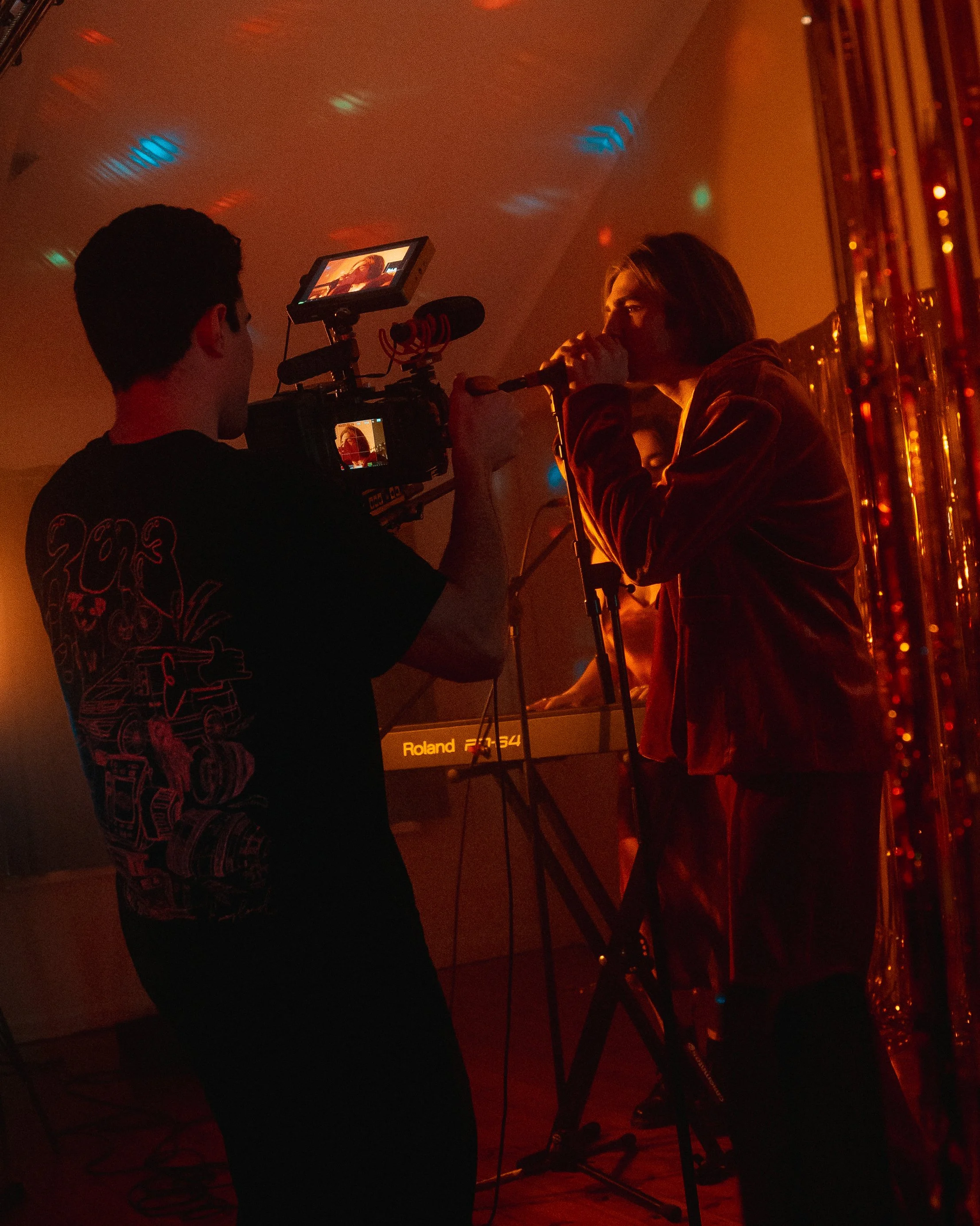A person is filming a woman singing into a microphone with a video camera, in a dimly lit, colorful venue with musical equipment and reflective decorations.