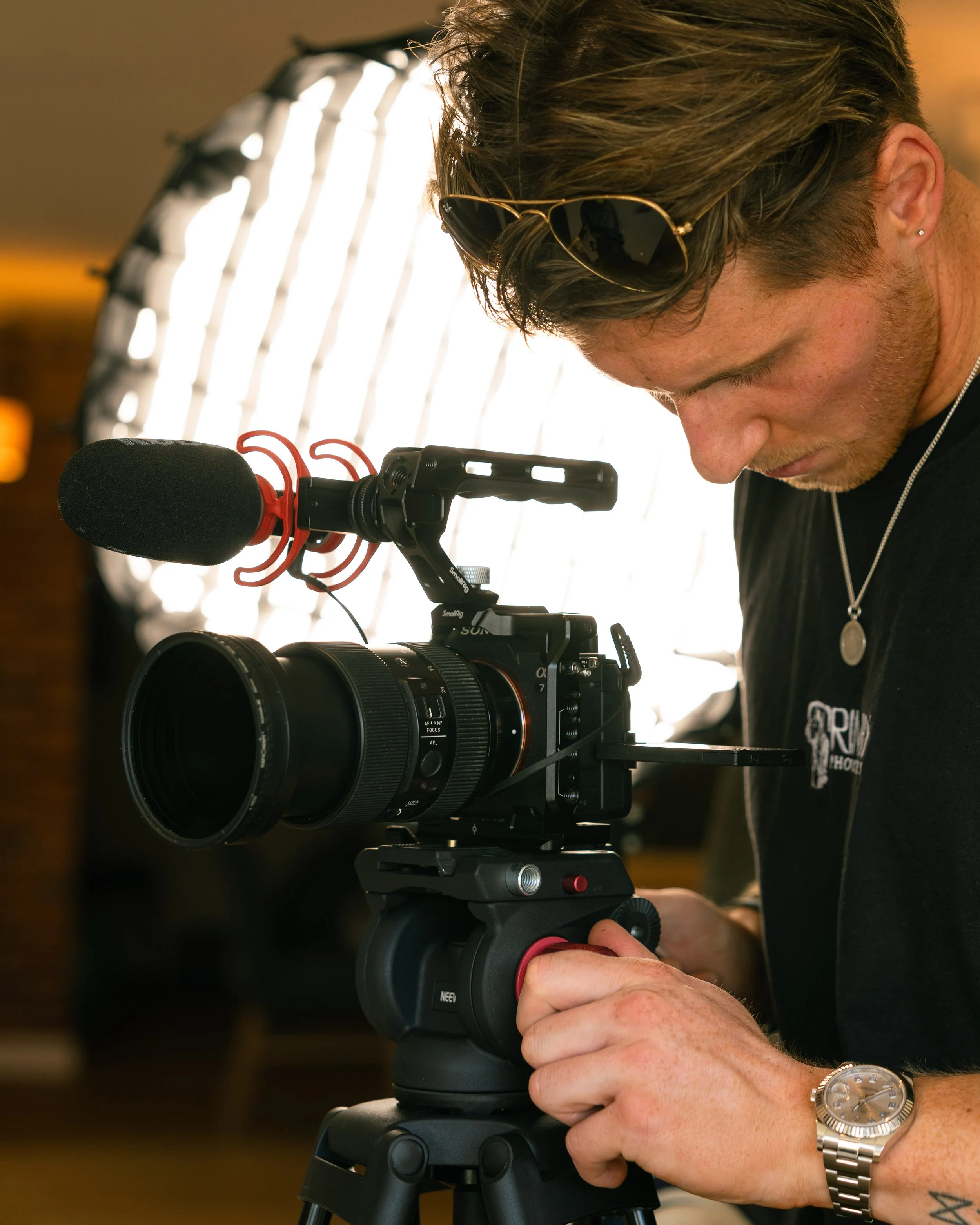 A man operating a professional video camera on a tripod, with a large light in the background.