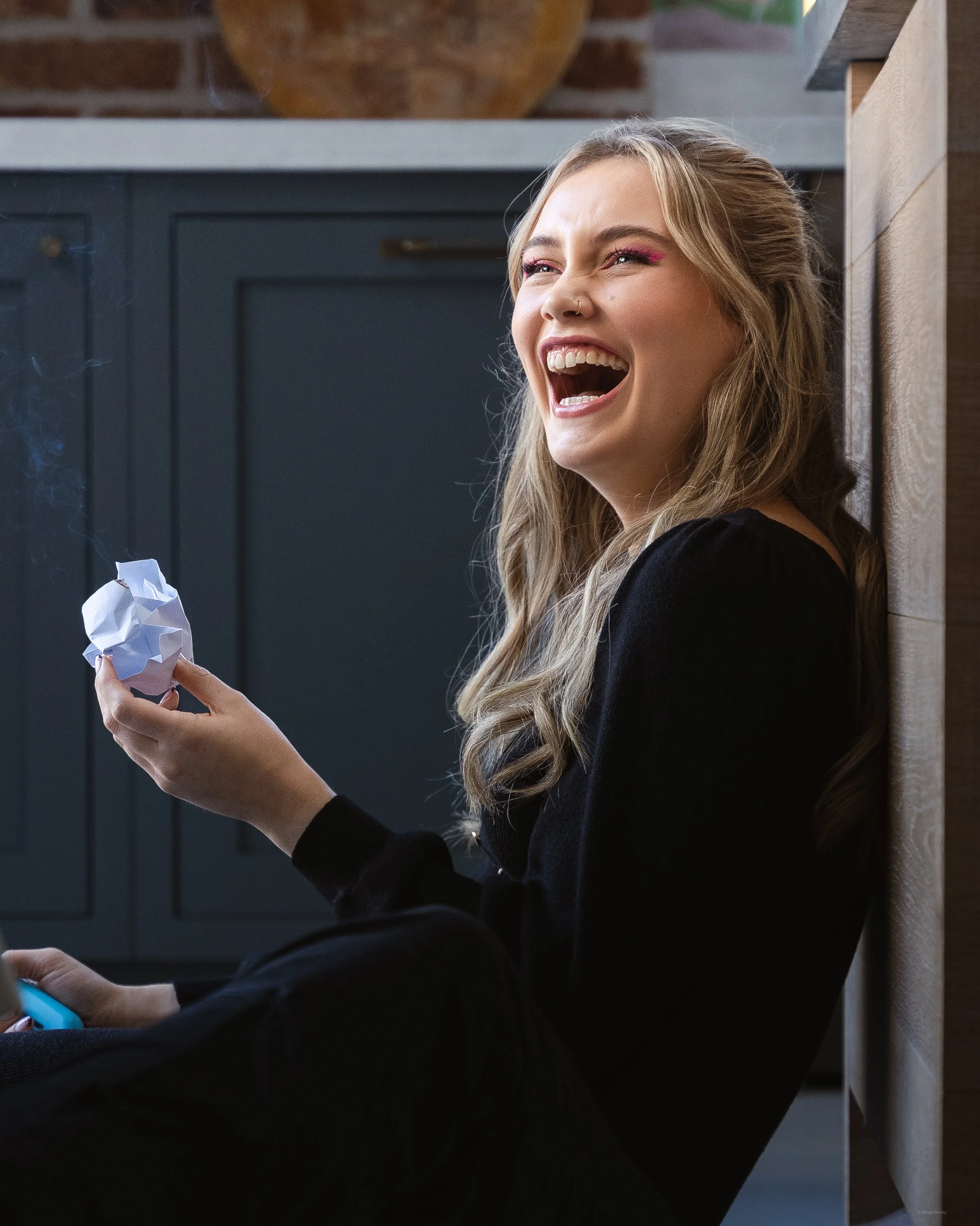 A young woman with long blonde hair and pink makeup is sitting on the floor, leaning against a wall, and laughing while holding crumpled paper.