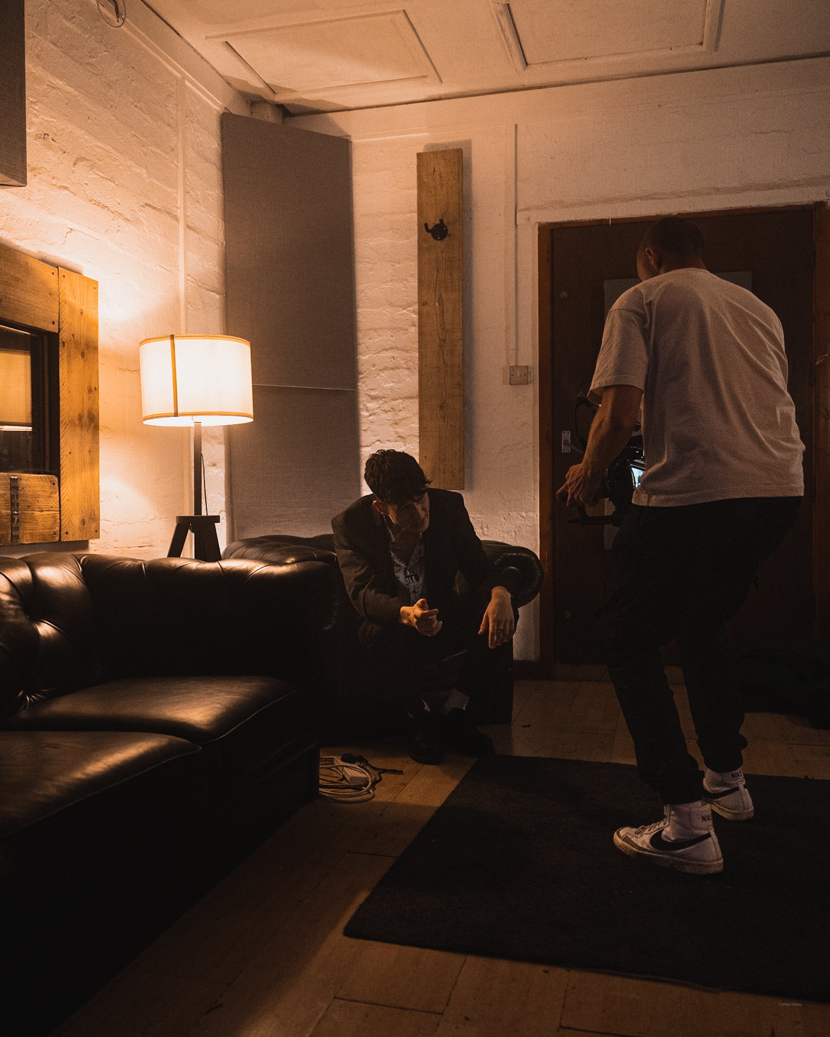 Two men are in a dimly lit room, one sitting on a black leather sofa talking on a cellphone, the other standing, adjusting a camera. The room has a lamp on a side table, white brick walls, and wooden accents.