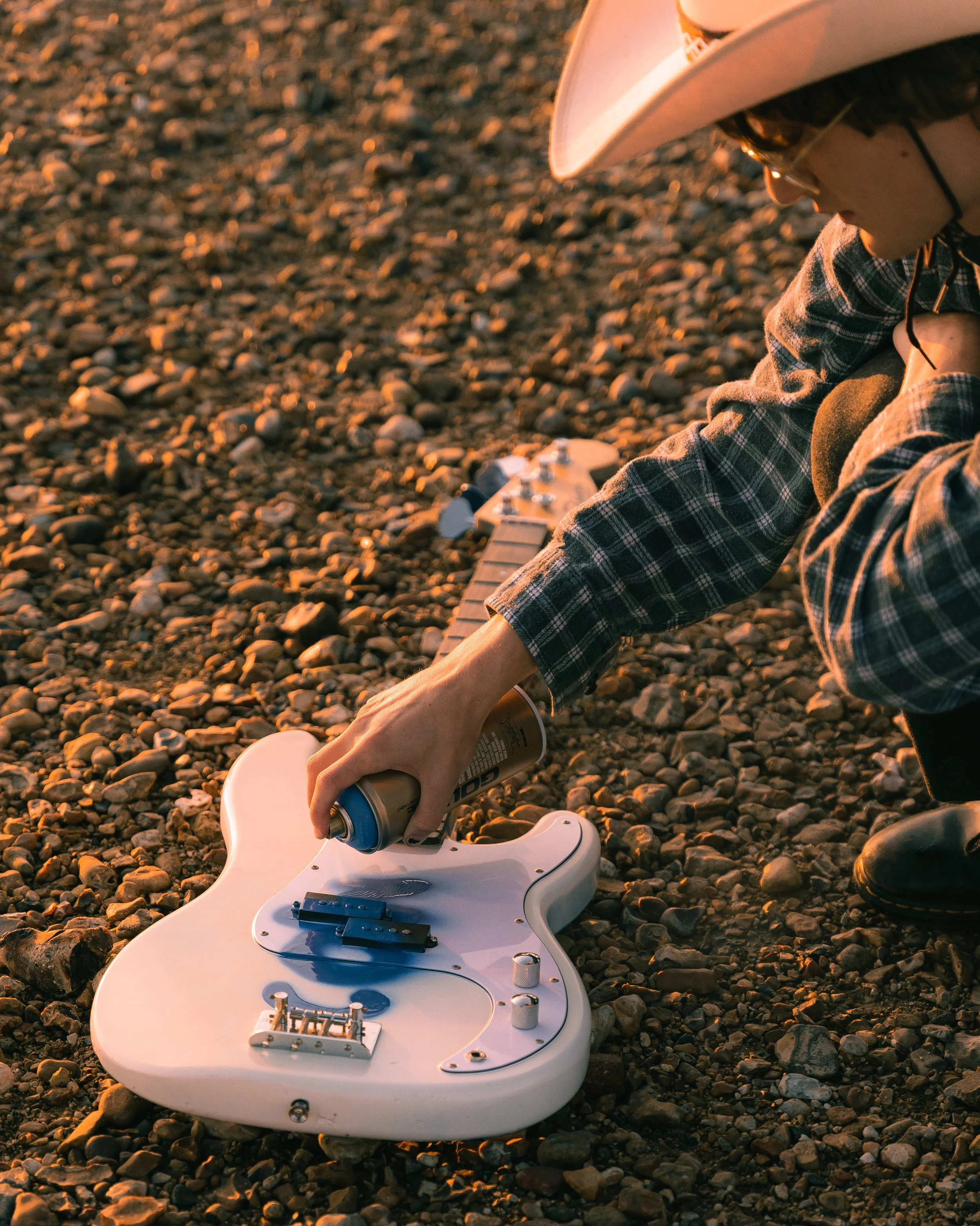 Person in a plaid shirt and cowboy hat spray painting a white electric guitar on rocky ground at sunset.