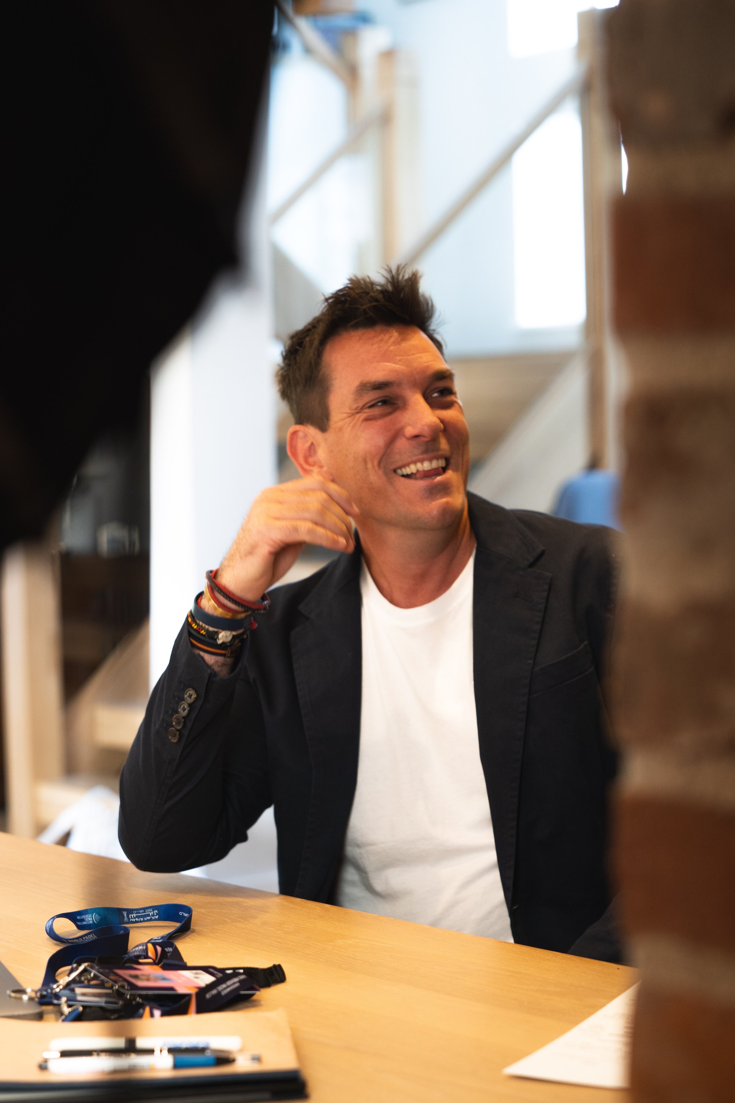 Man with dark hair smiling, seated at a desk, wearing a white T-shirt and dark blazer, with pens and lanyards on the desk.