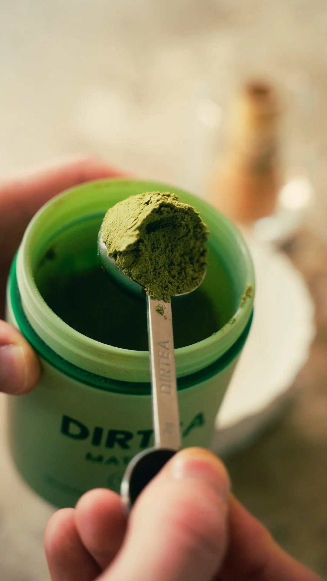 A spoonful of green powdered supplement labeled 'DIRTEA' being held over an open green container of the same supplement.