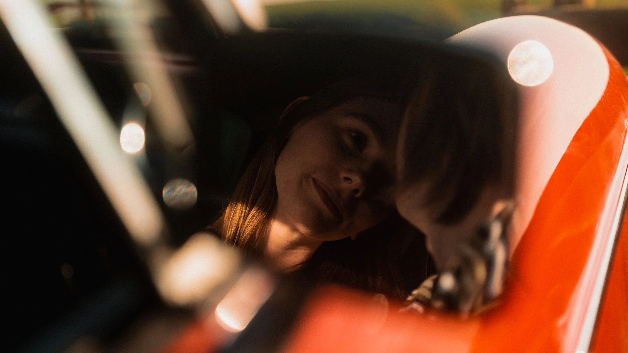 A woman with long hair inside a car, looking through the window with a smile. The photo is taken from outside, focusing on her face through the glass.