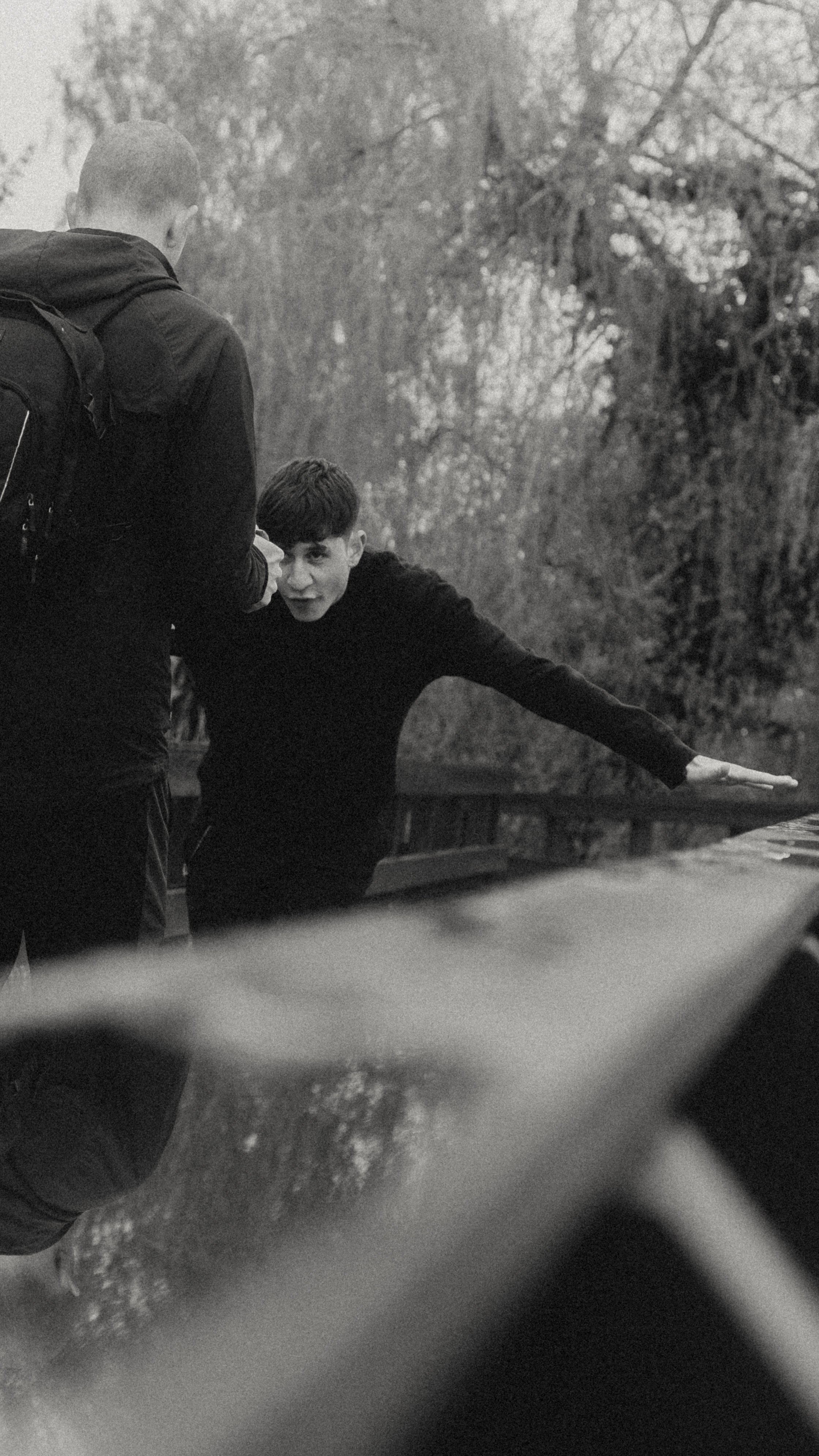 A young man with short hair and a dark sweater leans on a bridge railing, speaking to a person with short hair and a backpack, outdoors with trees in the background.