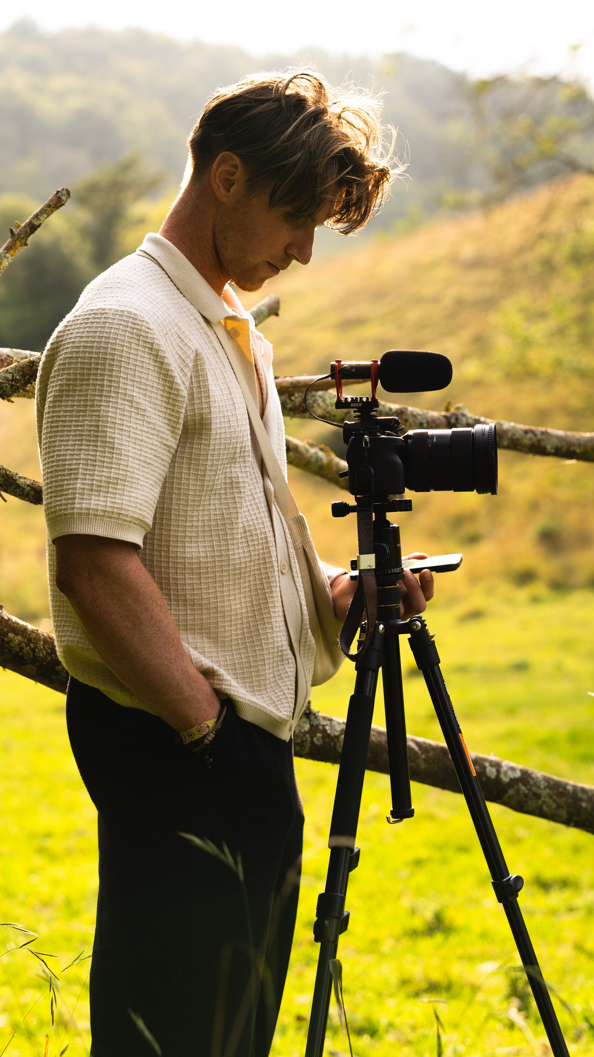 A man using a camera on a tripod outdoors during daytime, with sunlight and greenery in the background.