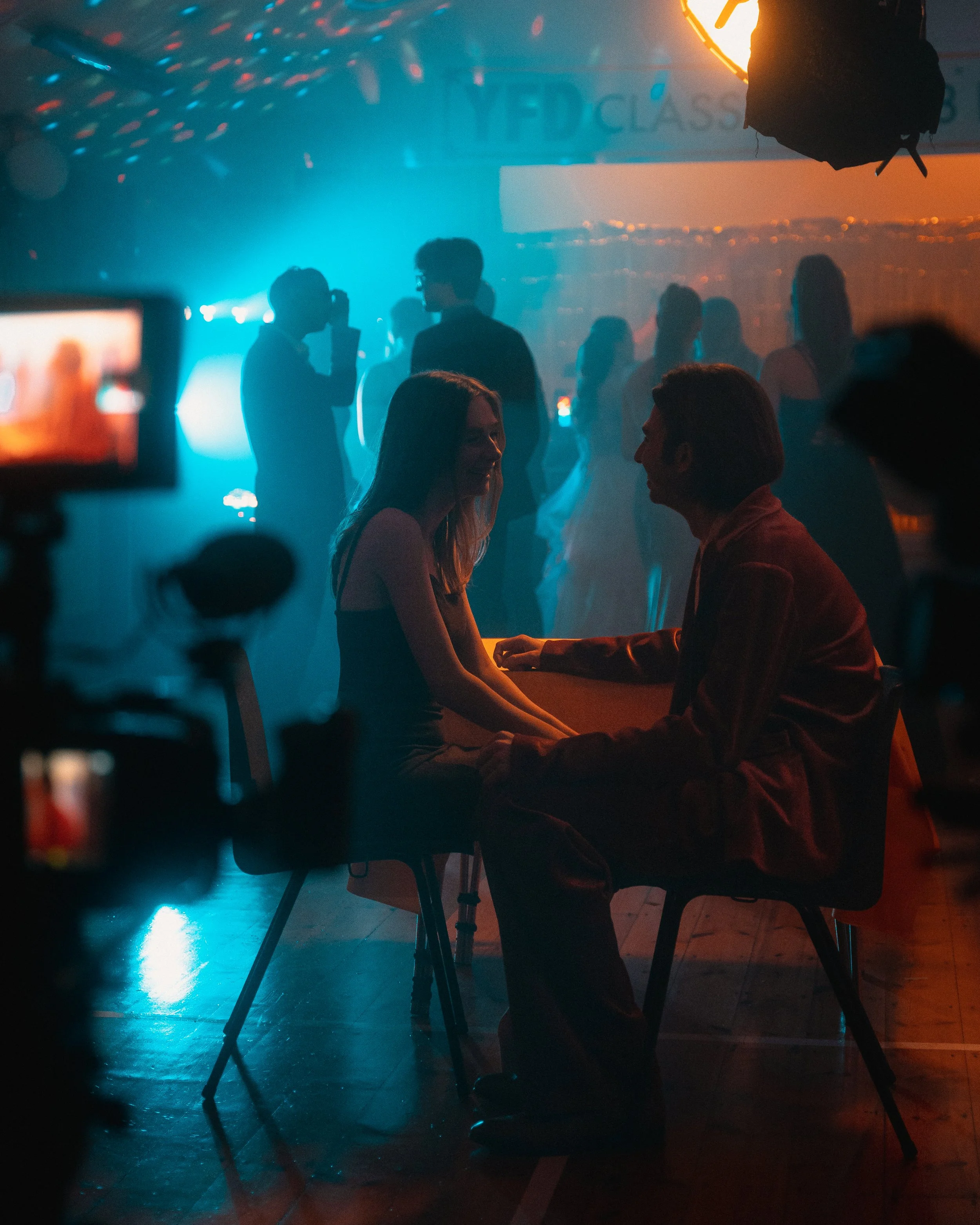 A dimly lit dance floor with people dancing in the background and a couple sitting and talking at a table in the foreground, illuminated by colorful lights.