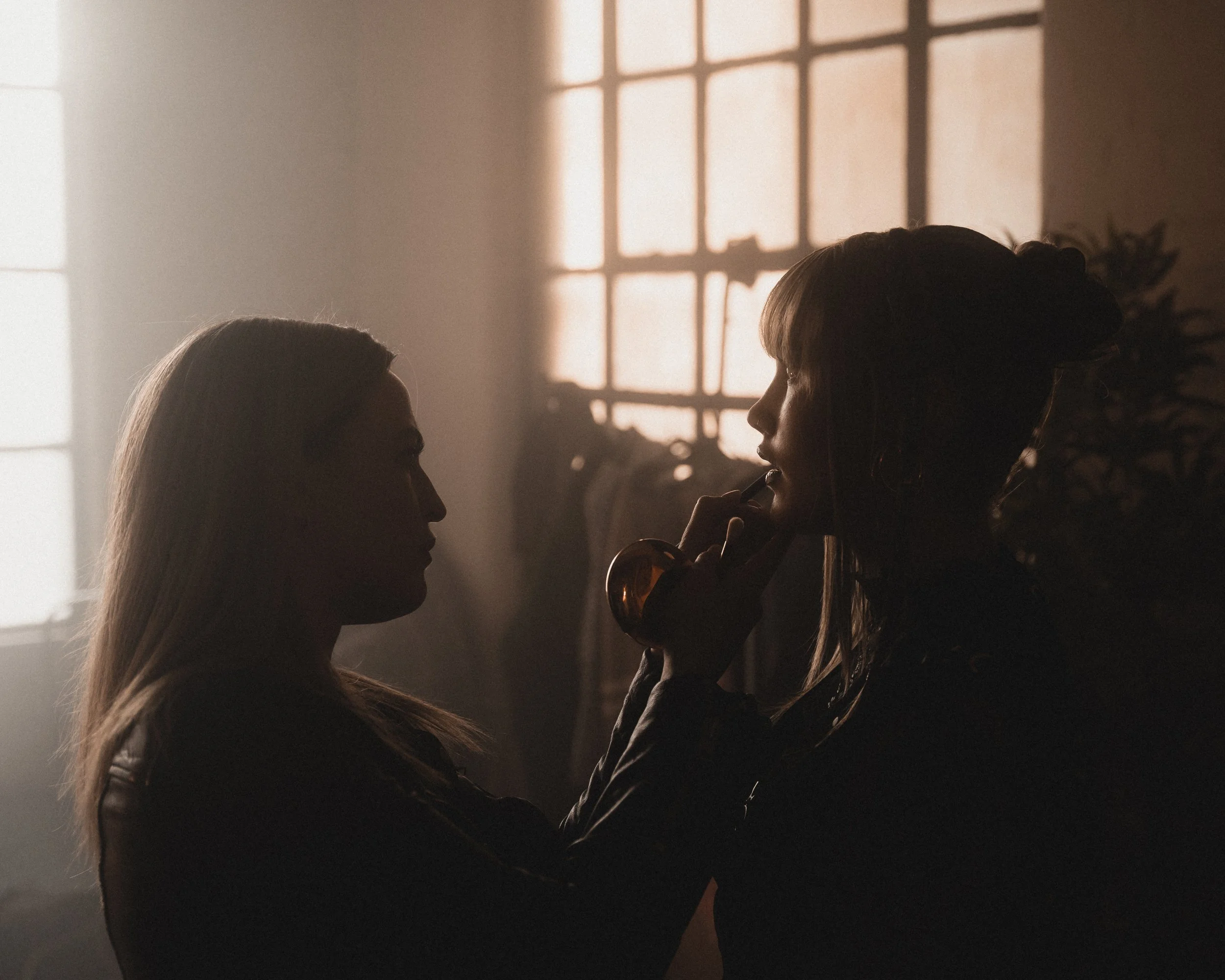 Two women in a dimly lit room with backlit windows facing each other, one woman applying lipstick to the other.