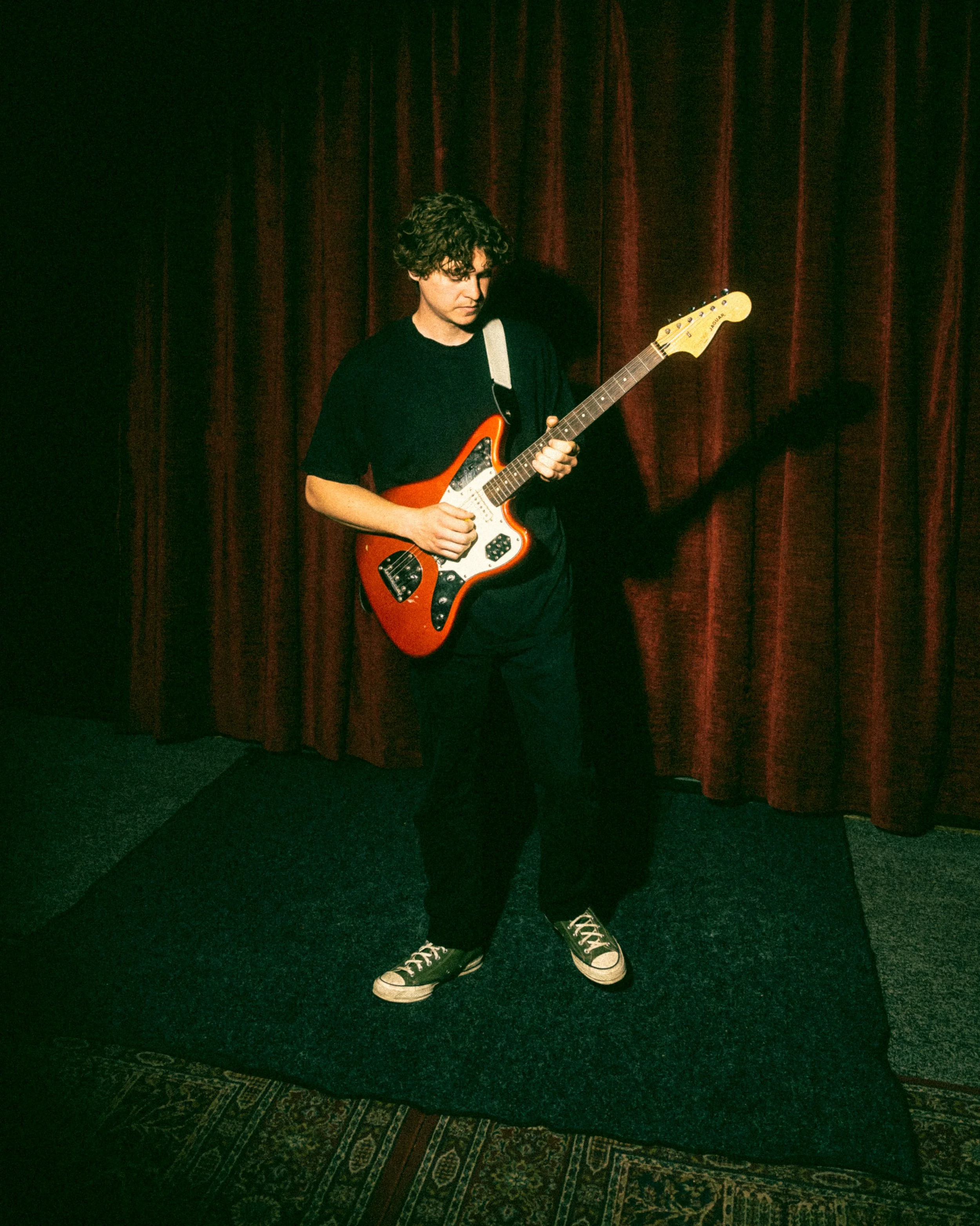 A young man with curly hair playing an electric guitar on a small stage in front of red velvet curtains.