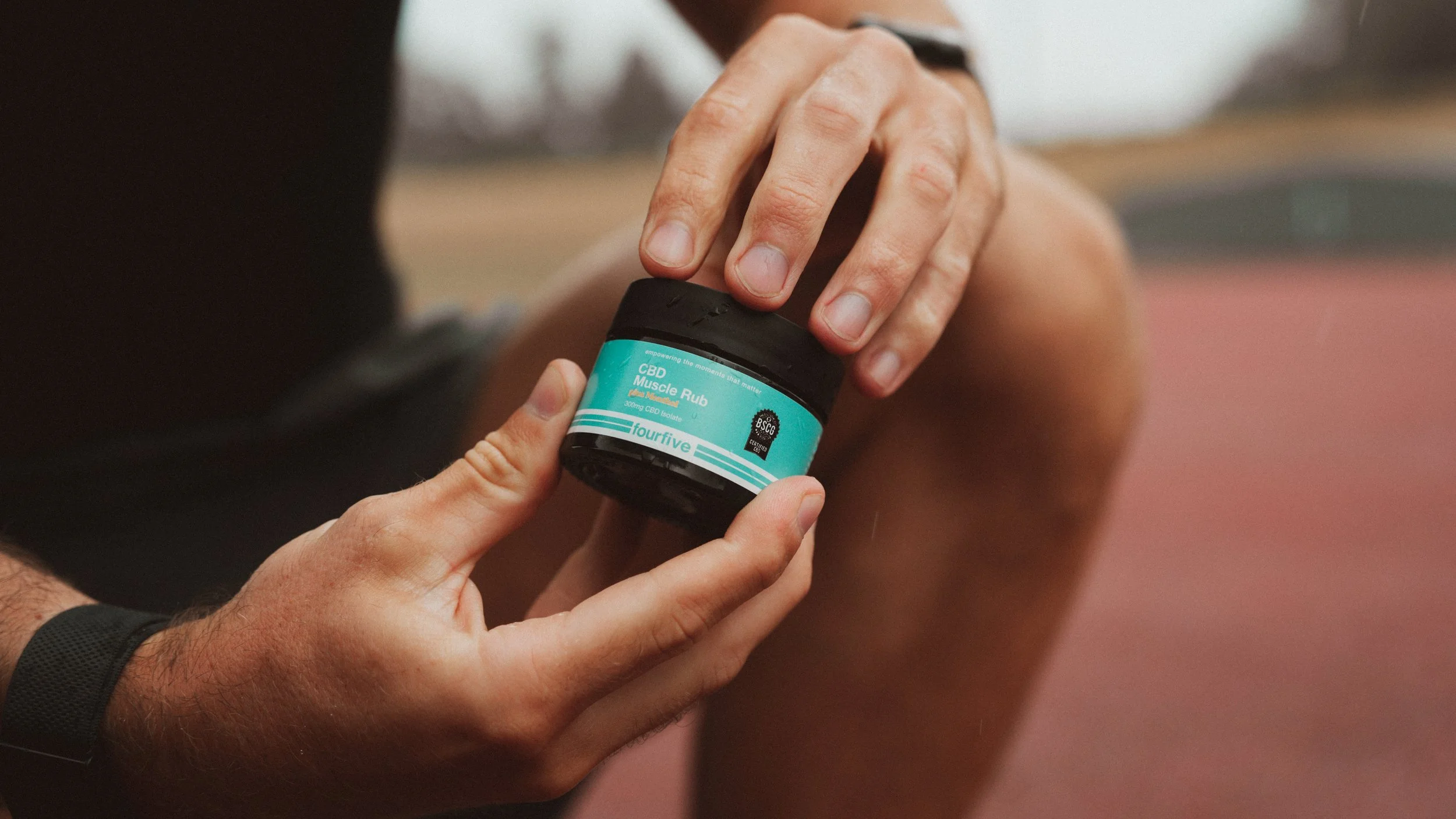 A person applying CBD muscle rub from a small jar labeled 'CBD Muscle Rub' with a teal label, at an outdoor track or sports field.