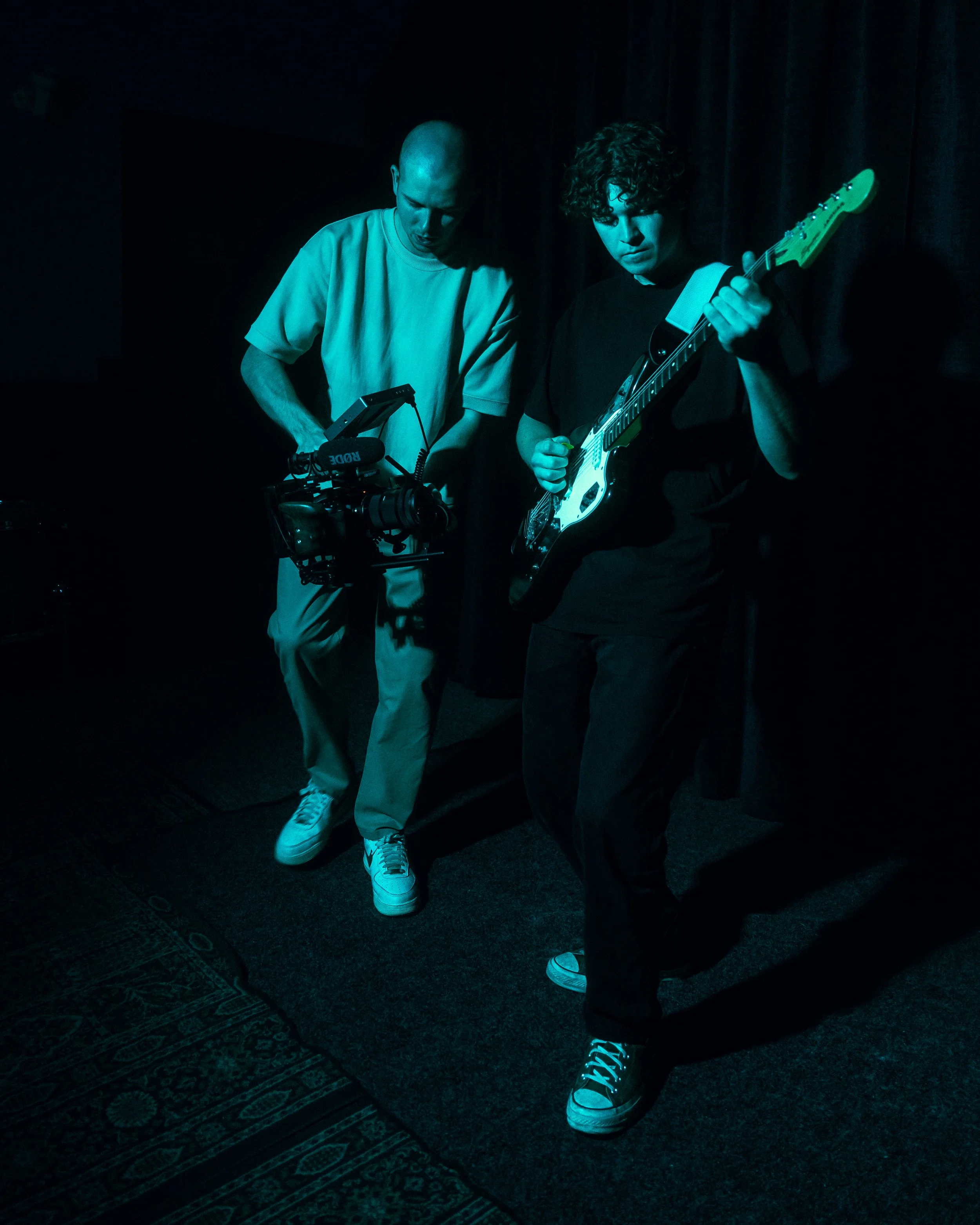 Two musicians, one playing an electric guitar and the other working with camera equipment, in a dimly lit setting with dark curtains in the background.