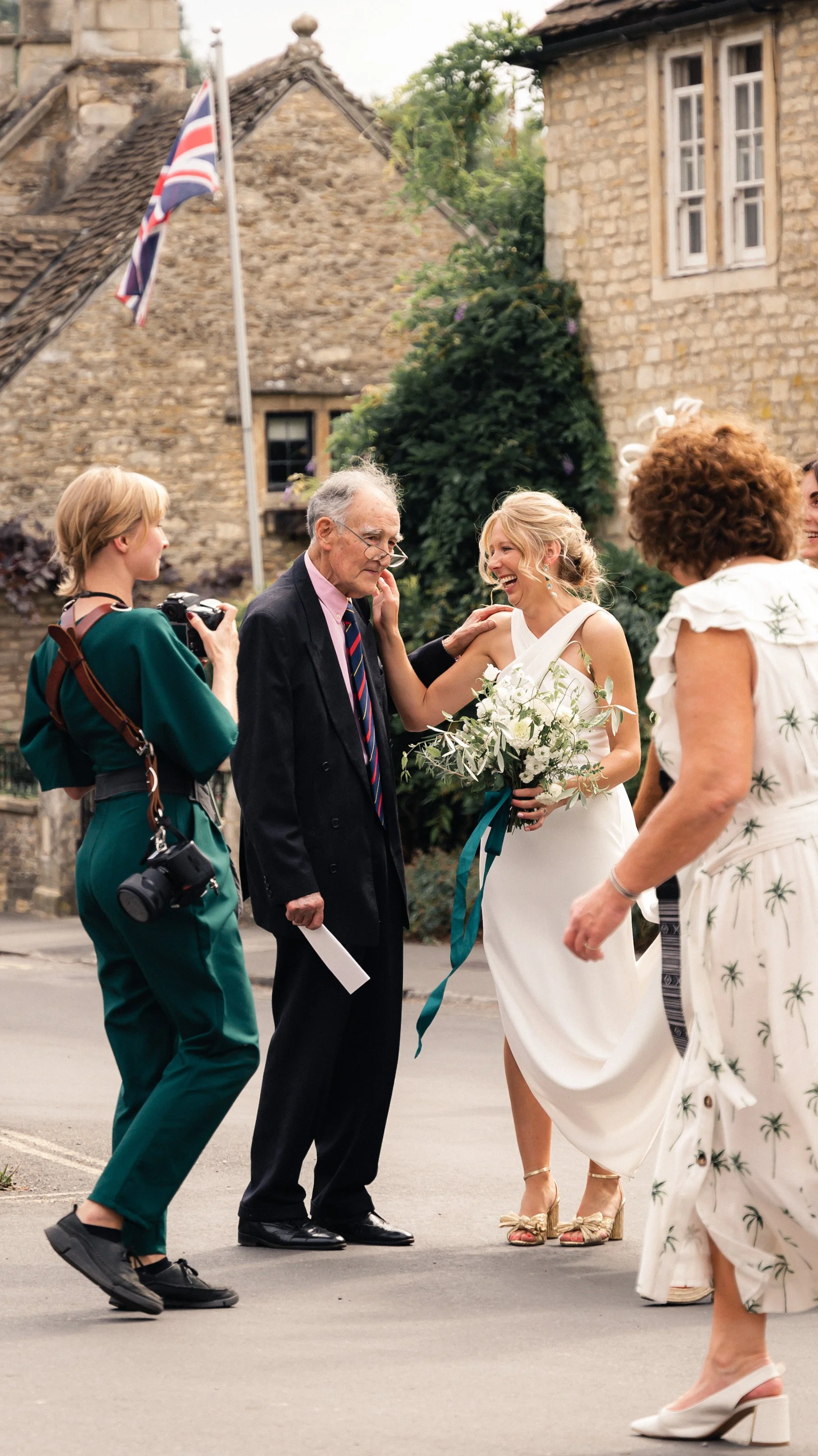 A bride in a white wedding dress holding a bouquet, smiling and looking at an older man in a suit, outside in front of stone buildings with a British flag, surrounded by women taking photos