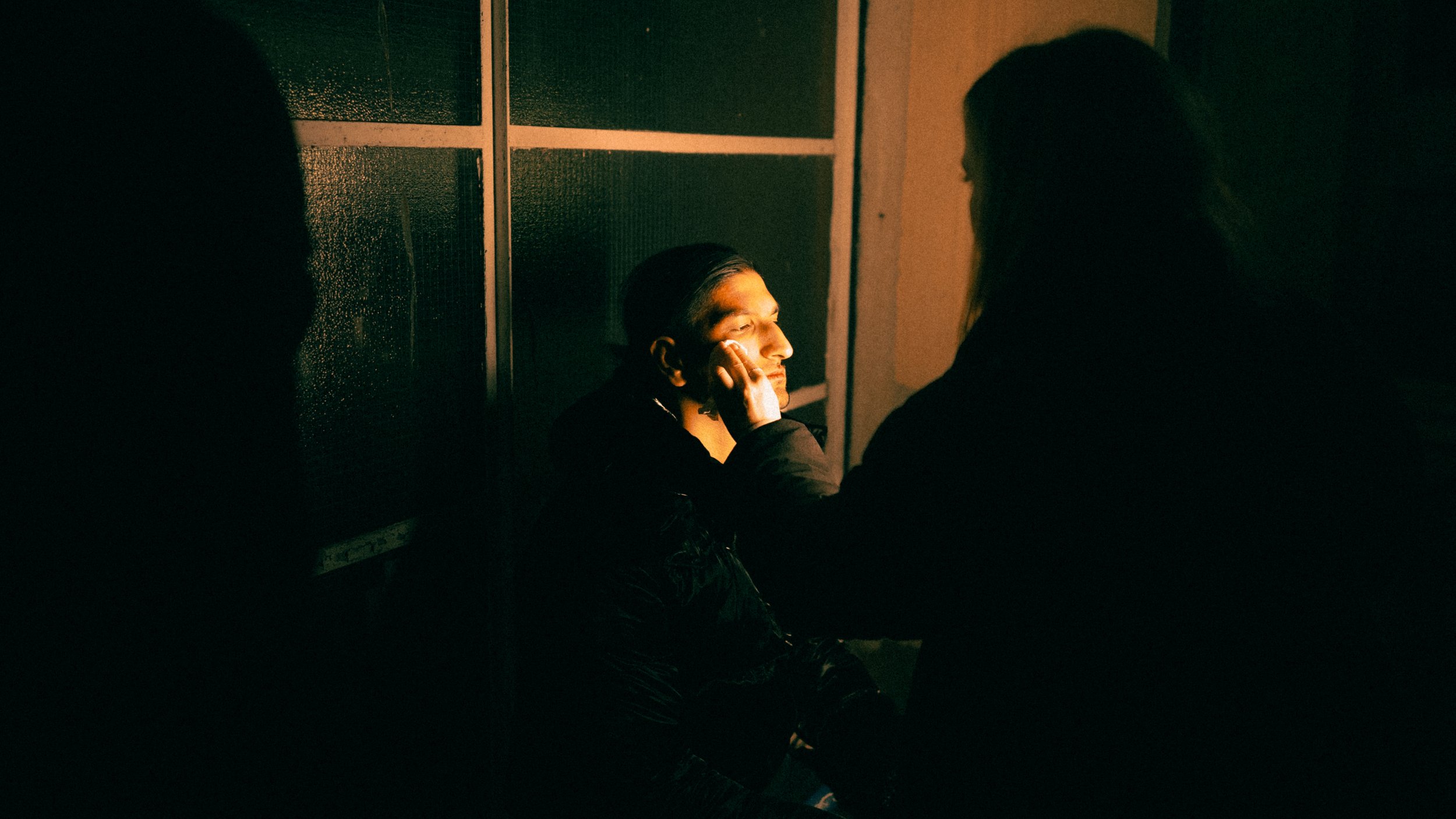 A person receives makeup application from another person in a dimly lit room. The person is seated with eyes closed, while the other person gently touches their face.
