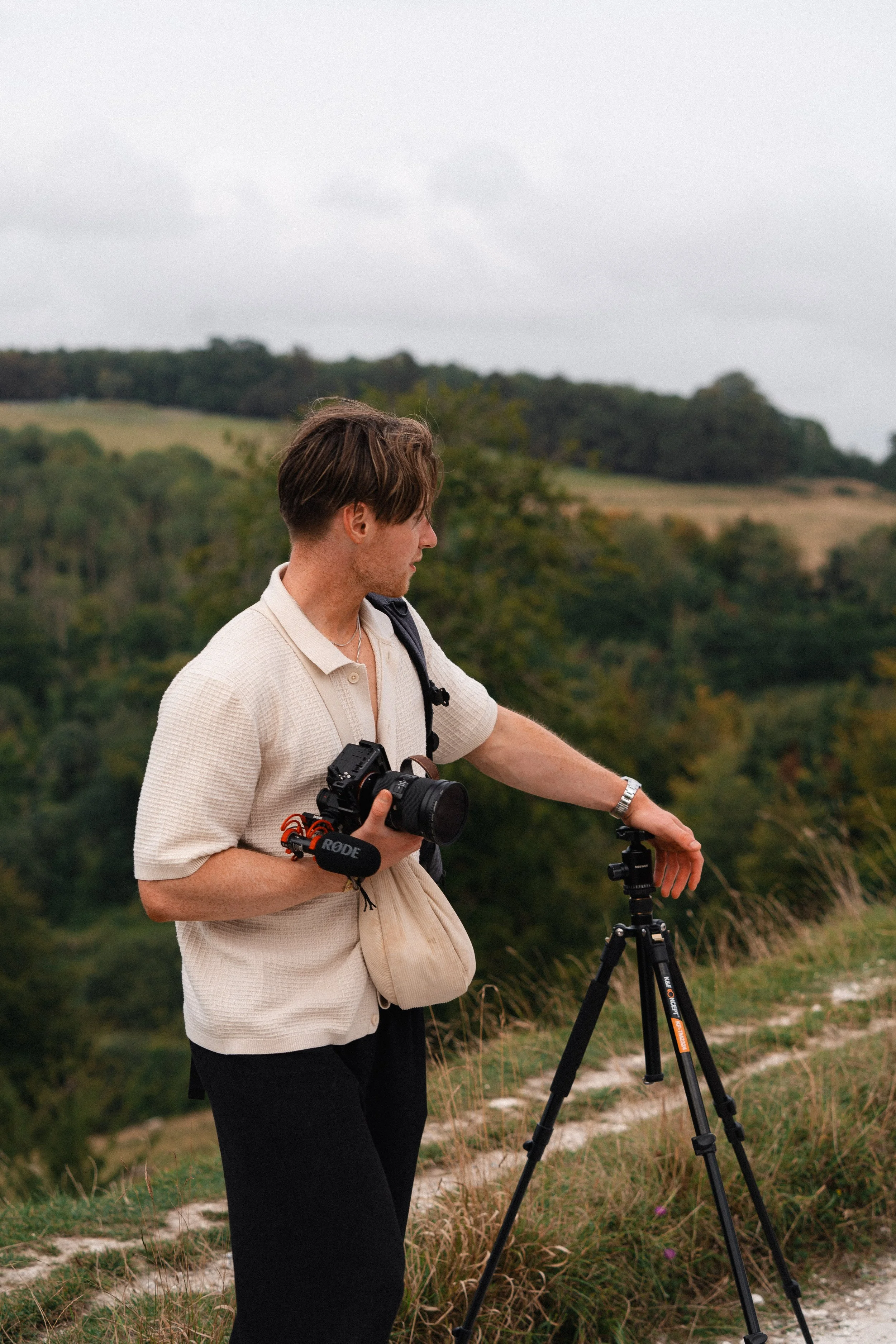 A person standing outdoors on a hillside with trees and rolling hills in the background, adjusting a camera on a tripod.