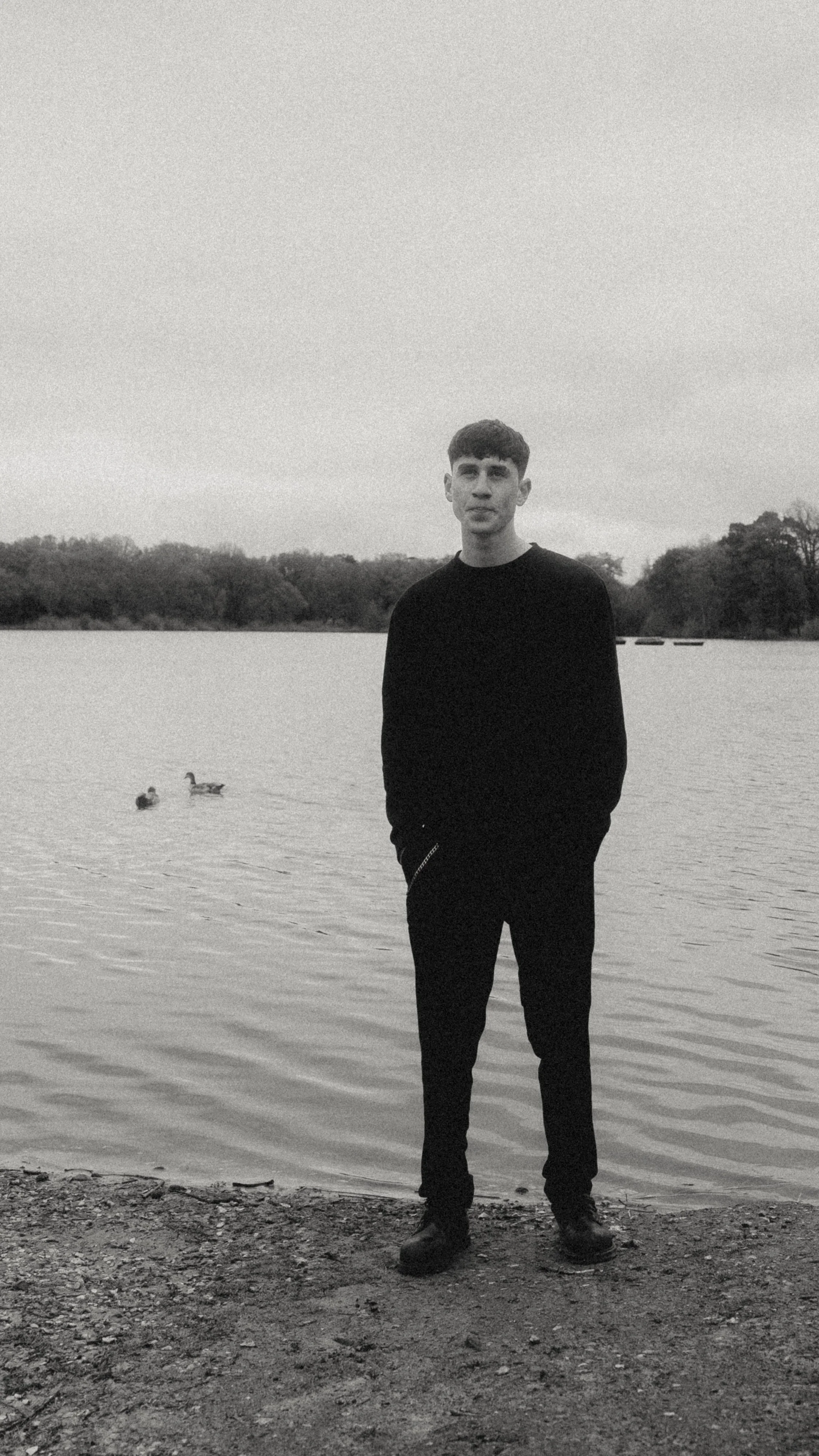 A young man stands on a lakeshore with two ducks swimming behind him in a black and white photo.