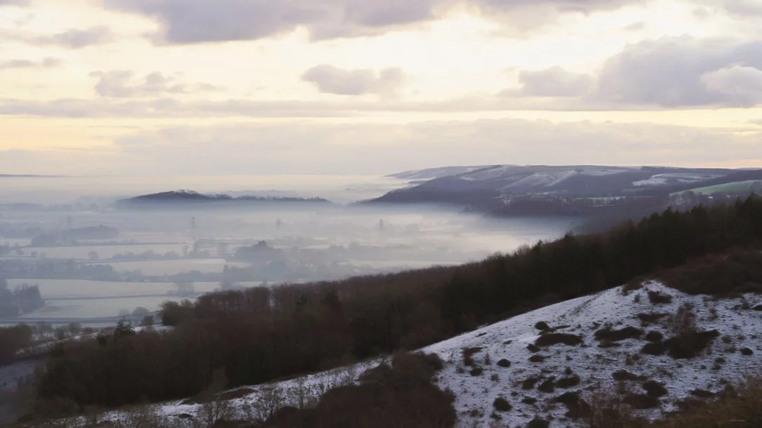 Landscape view of rolling hills with some snow, trees, and a misty valley under a cloudy sky