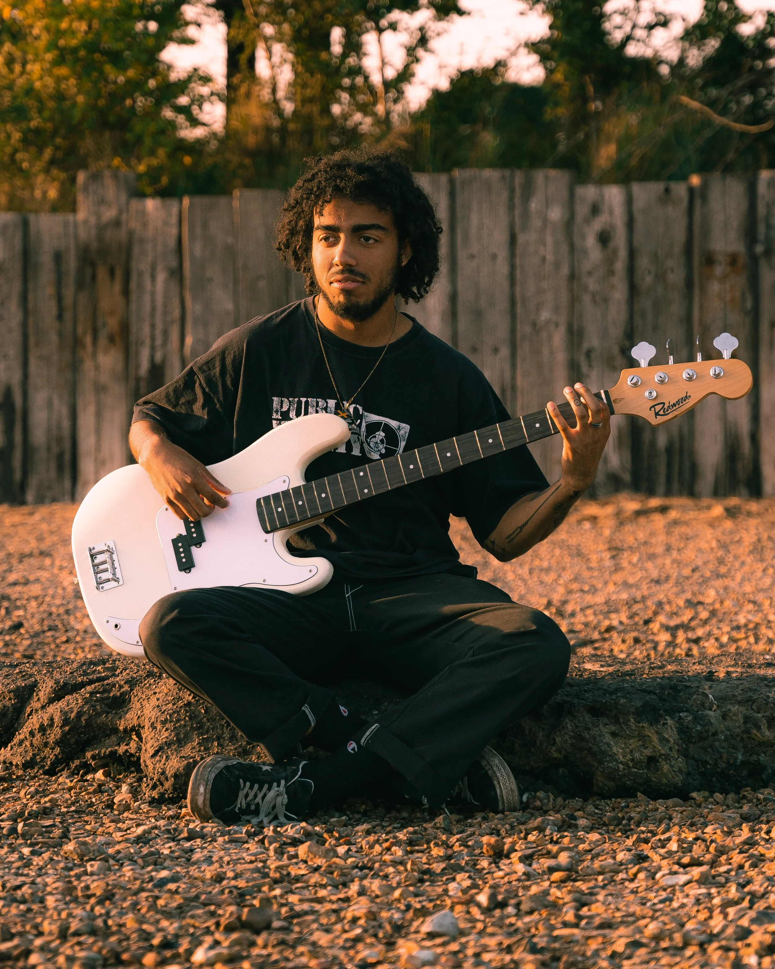 A young man with curly hair and a beard sitting on rocks outdoors, playing a white electric guitar, with a wooden fence and trees in the background during sunset.