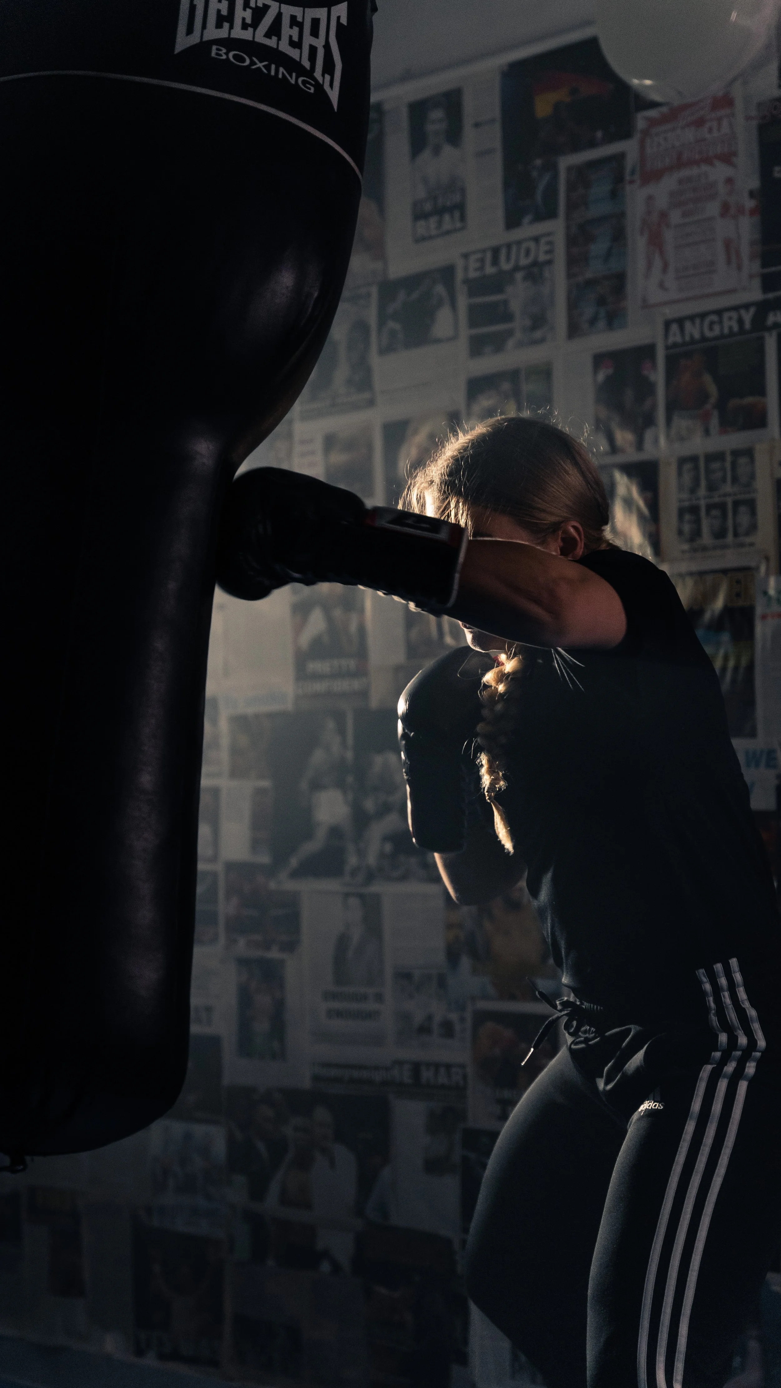 A woman wearing black athletic clothes and gloves is practicing boxing on a punching bag in a gym. The gym has walls decorated with various posters and images.