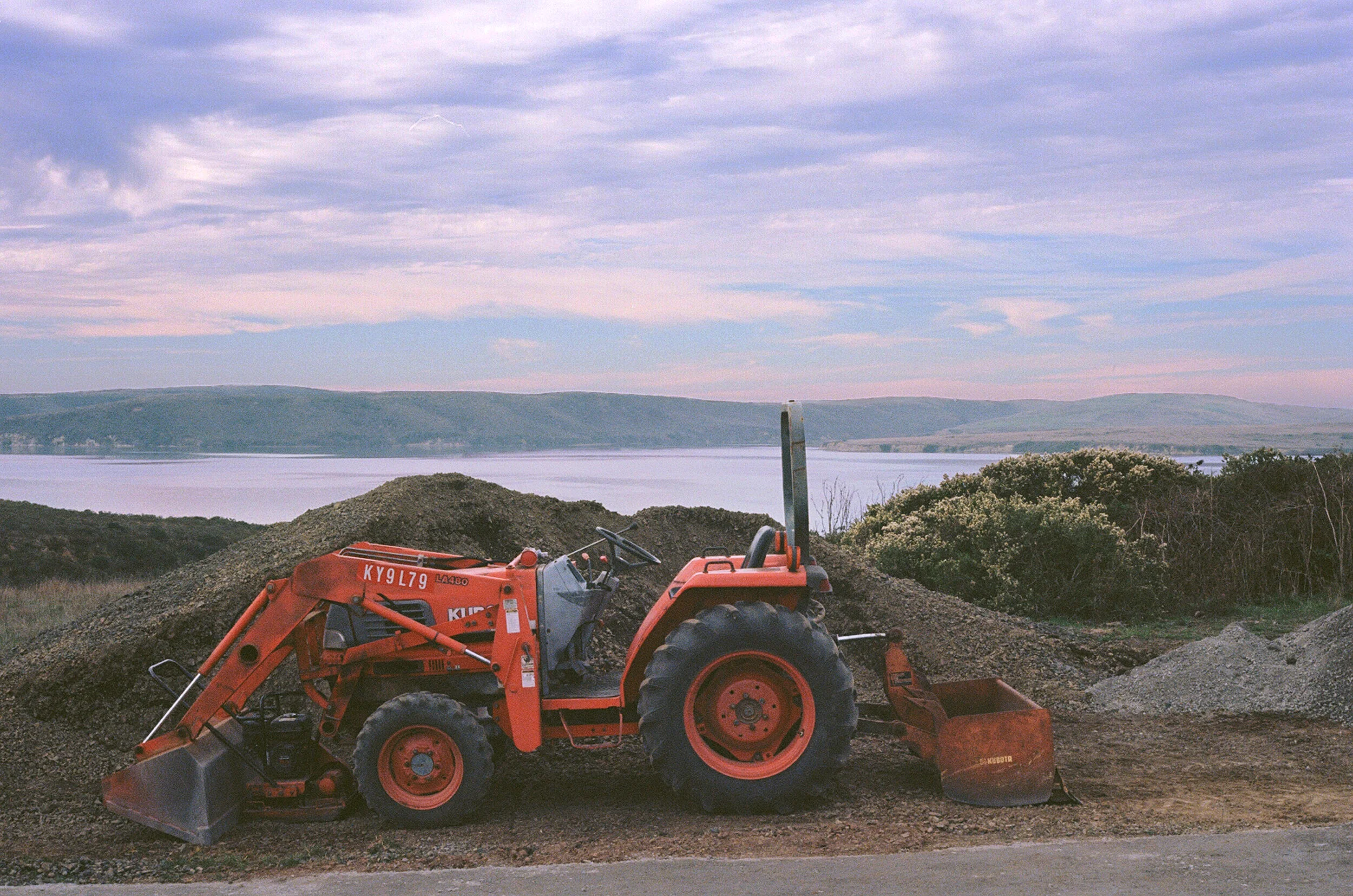 A small tractor rests on the edge of the Estero Trail, the Estero and some coyote bush glowing purple in the last of the day's sunlight.