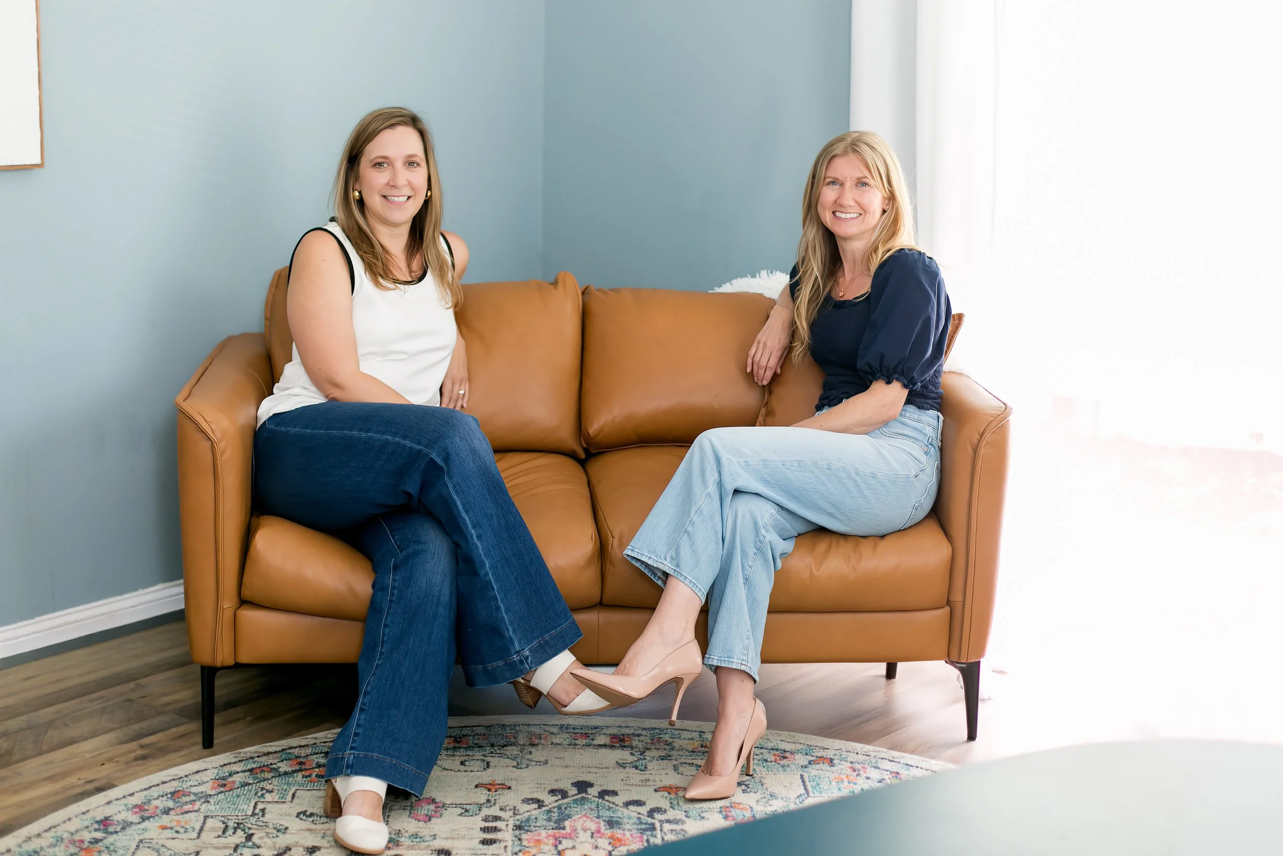 A female therapist sitting on a tan leather sofa in a brightly lit room with blue walls and white curtains, listening to her client speak.