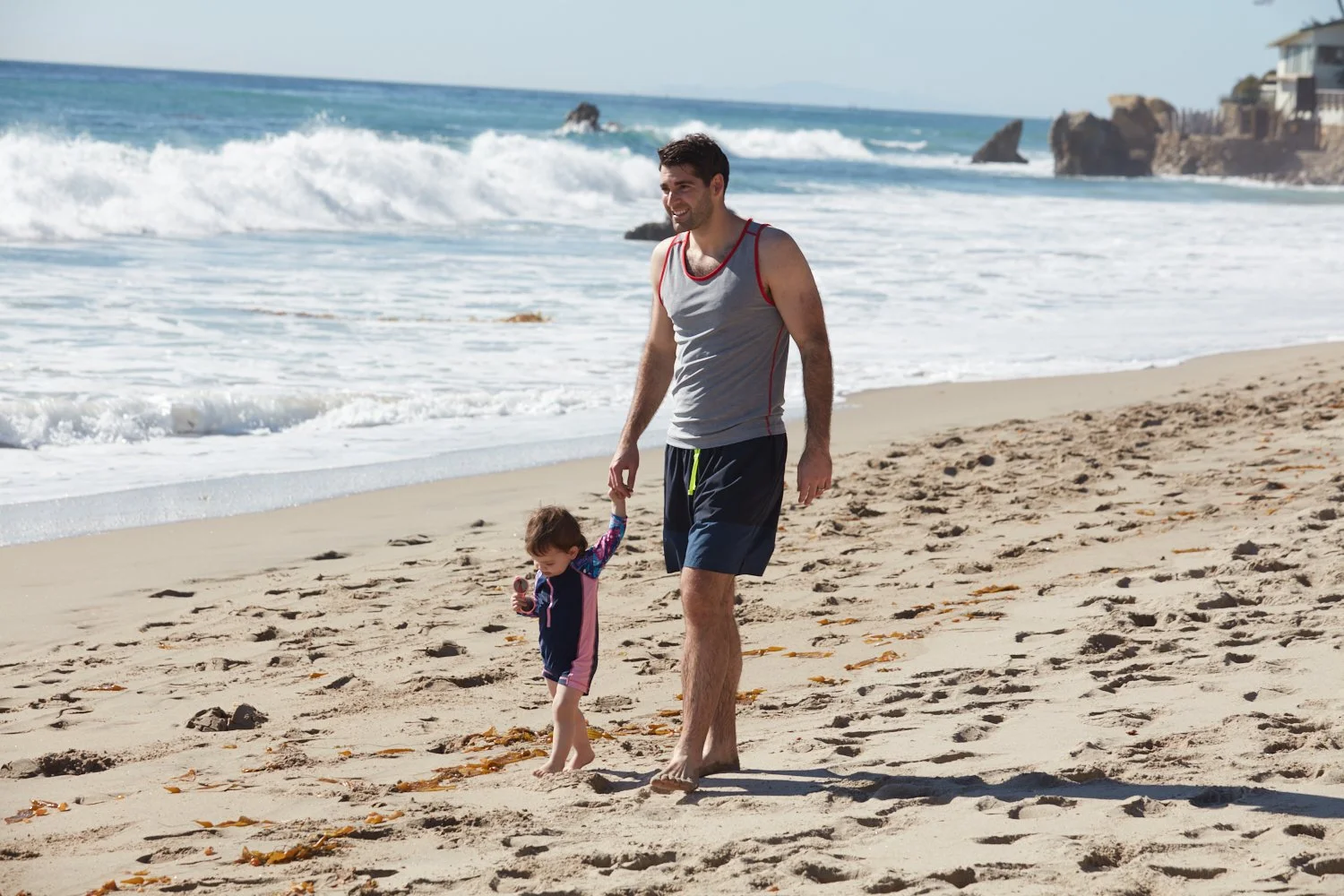 A man and a young girl walking hand in hand on a sandy beach near the ocean.