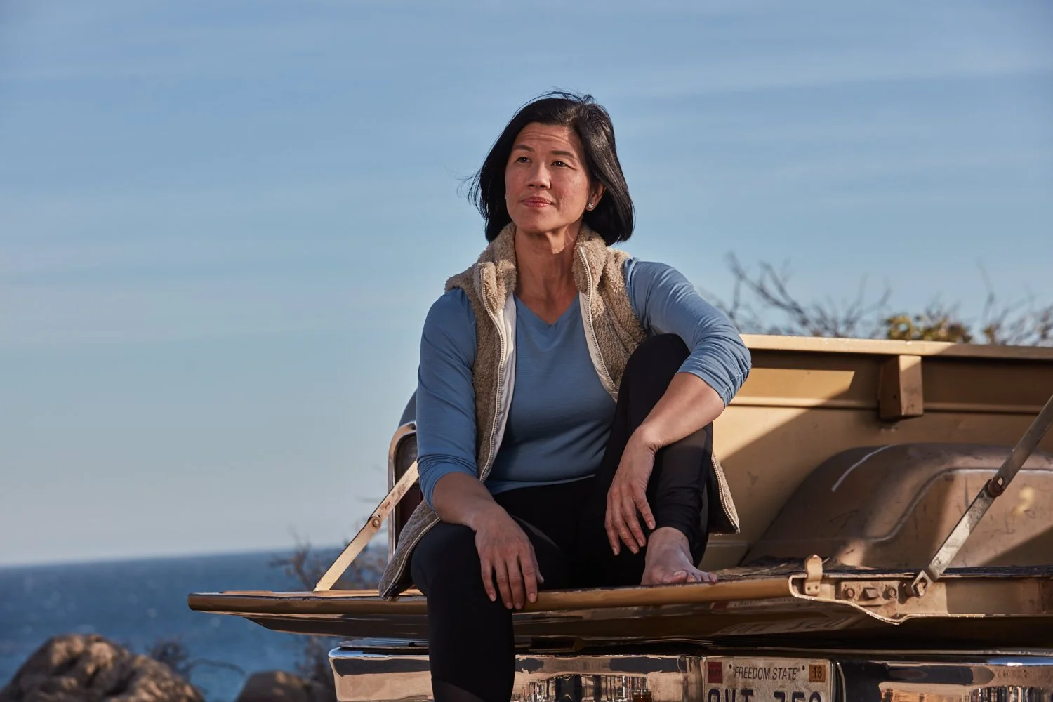 A woman sitting on the tailgate of a truck near the ocean with rocky shoreline in the background, wearing a blue shirt and a fleece vest.