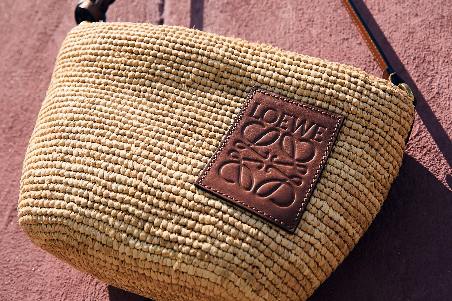 A woven straw handbag with a leather patch that has the Loewe logo embossed on it, placed on a reddish surface.