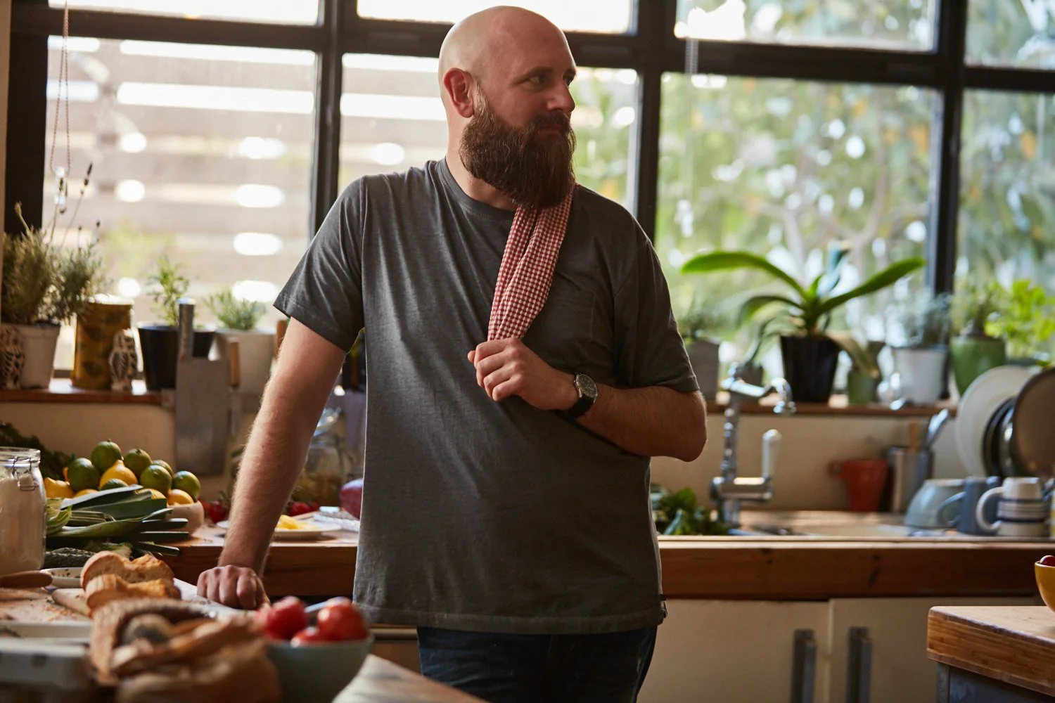 A bearded man in a gray t-shirt with a checkered towel over his shoulder, standing in a bright kitchen surrounded by fresh fruits, vegetables, and plants.