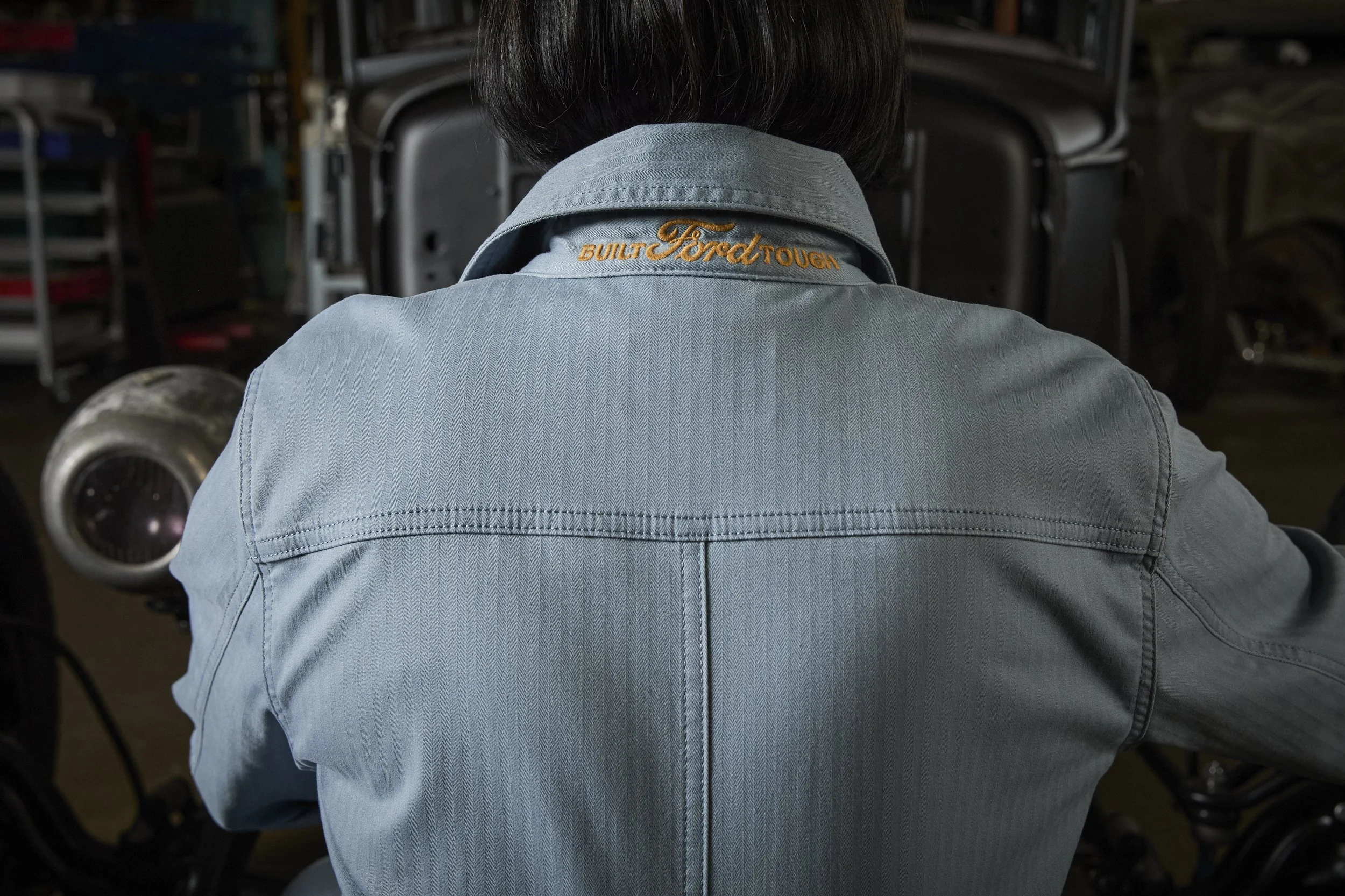 Back of a person wearing a light blue denim shirt with the words "Built Ford Tough" embroidered on the collar, sitting in front of an industrial or workshop setting.