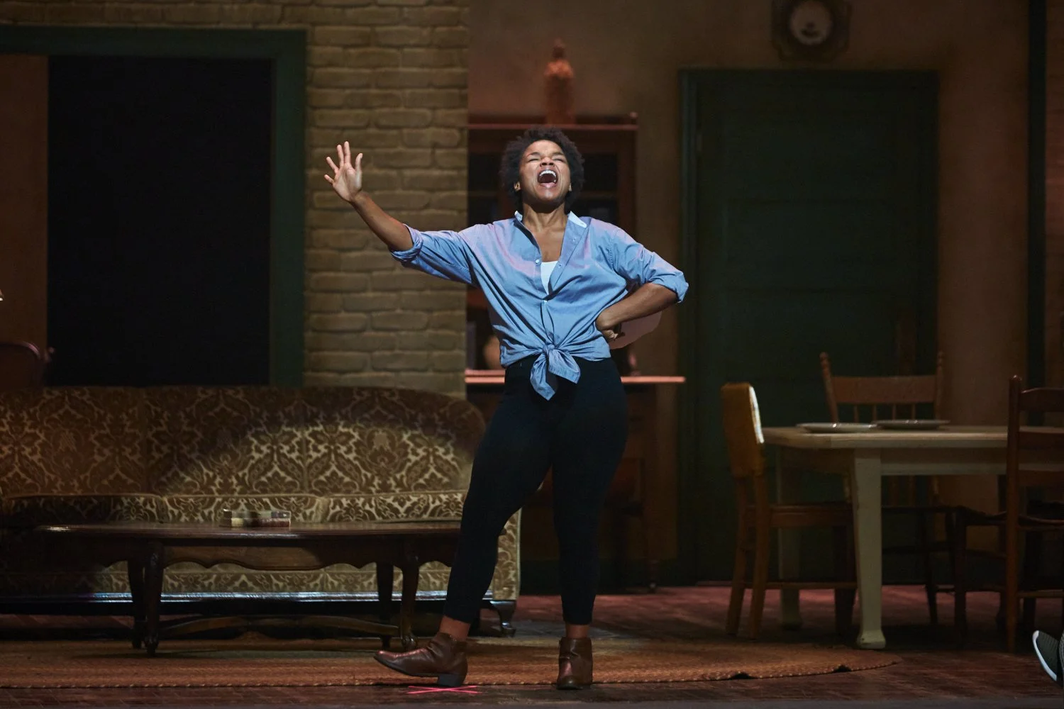 A woman standing on stage in a room, mid-performance, wearing a blue shirt tied at the waist, black pants, and brown shoes, with a lively and expressive pose.