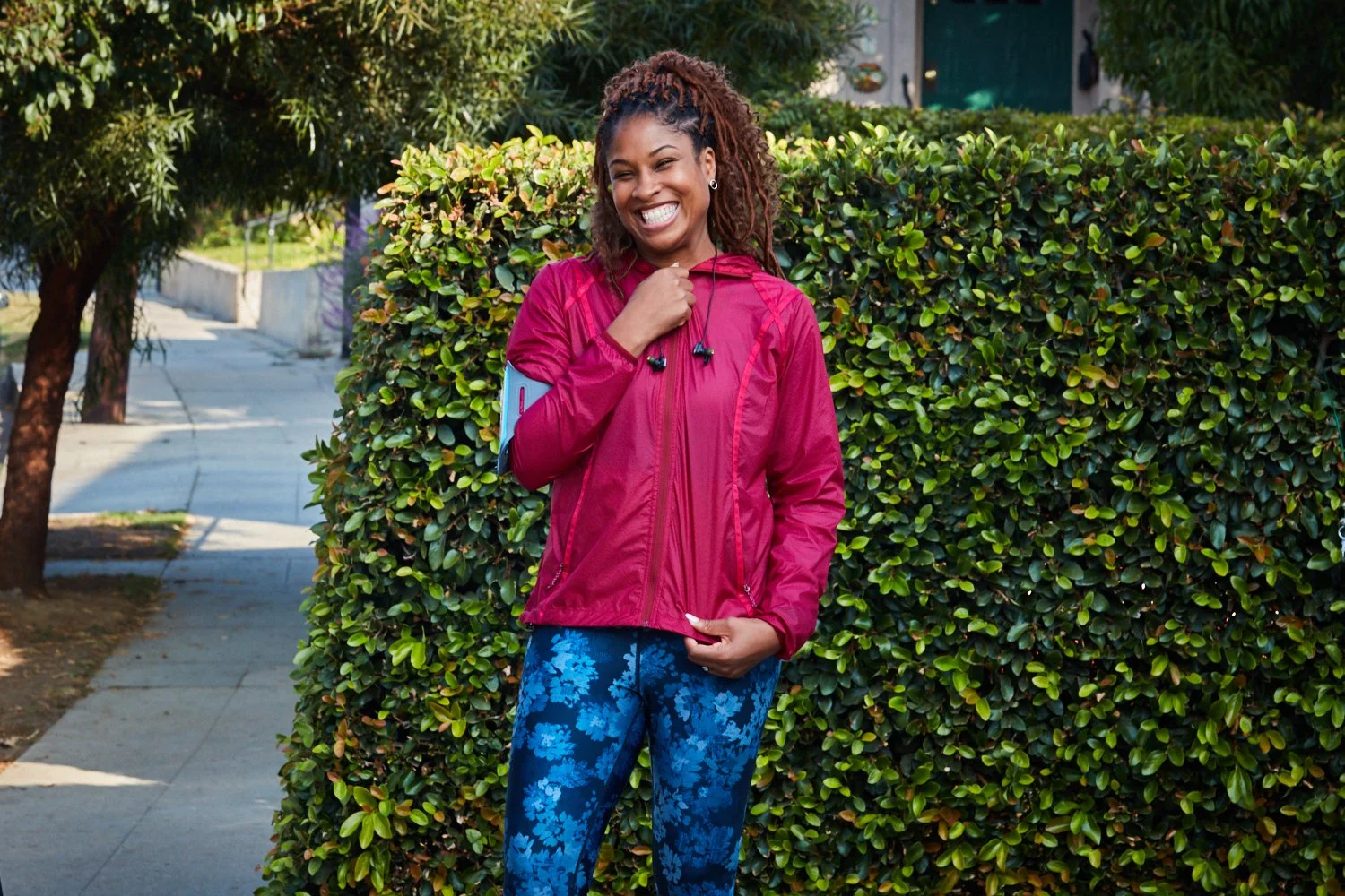 A woman smiling and standing outdoors on a sidewalk, wearing a pink jacket, blue floral leggings, and earphones, with a green hedge in the background.
