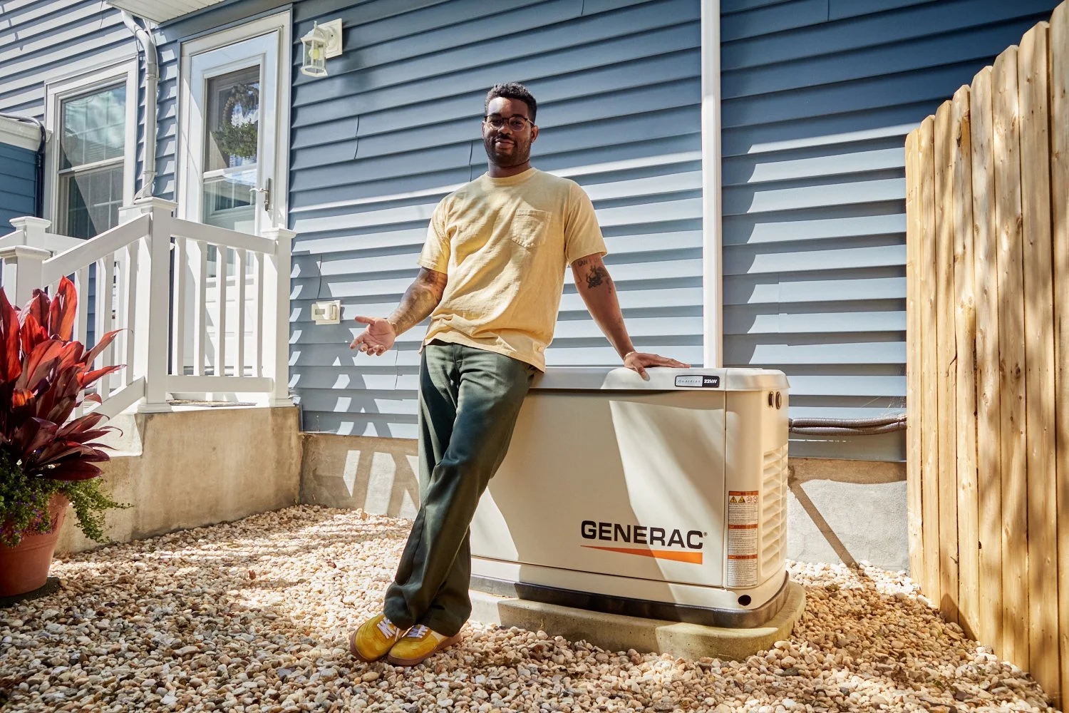 A man standing outdoors next to a Generac generator unit, gesturing with his right hand, in a backyard with a blue house exterior, a small staircase, potted plants, and a wooden fence.