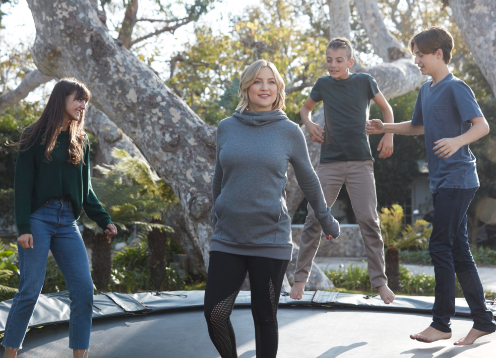 Four children and an adult woman stand around a trampoline outdoors, with the children jumping and the woman smiling.