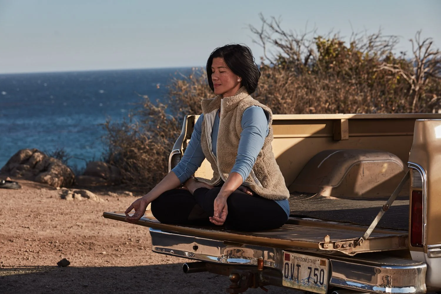 A woman practicing yoga in a seated meditative pose on the tailgate of a truck near the coast with ocean and rocks in the background.