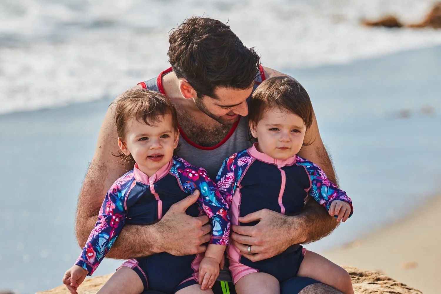 A man with two young children sitting on a beach, with ocean waves in the background.