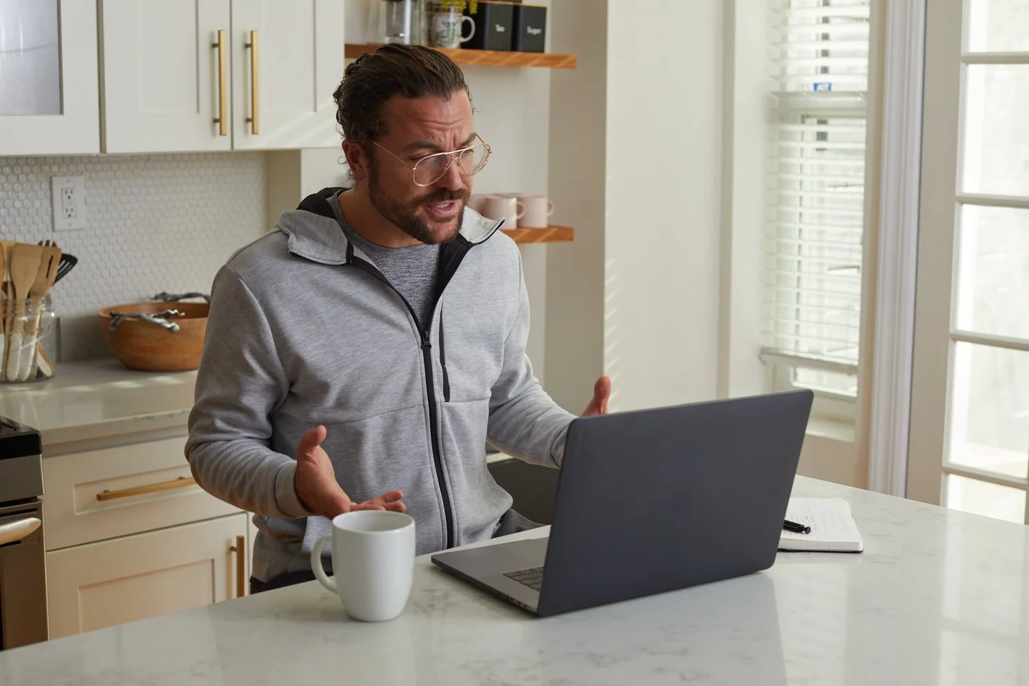 A man with glasses, a beard, and wearing a gray zip-up hoodie, appears to be speaking or explaining something while looking at his open laptop on a white kitchen countertop. There's a white coffee mug near the laptop. The background features white ca