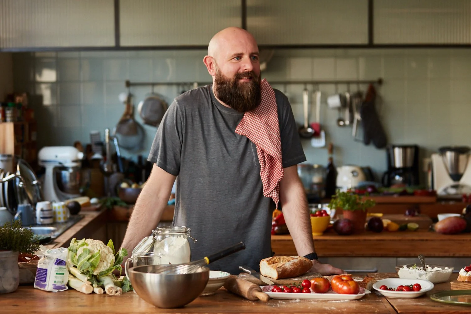 A bearded man in a gray t-shirt stands in a rustic kitchen with a red checkered towel on his shoulder, surrounded by fresh vegetables, bread, and cooking ingredients on the wooden countertop.
