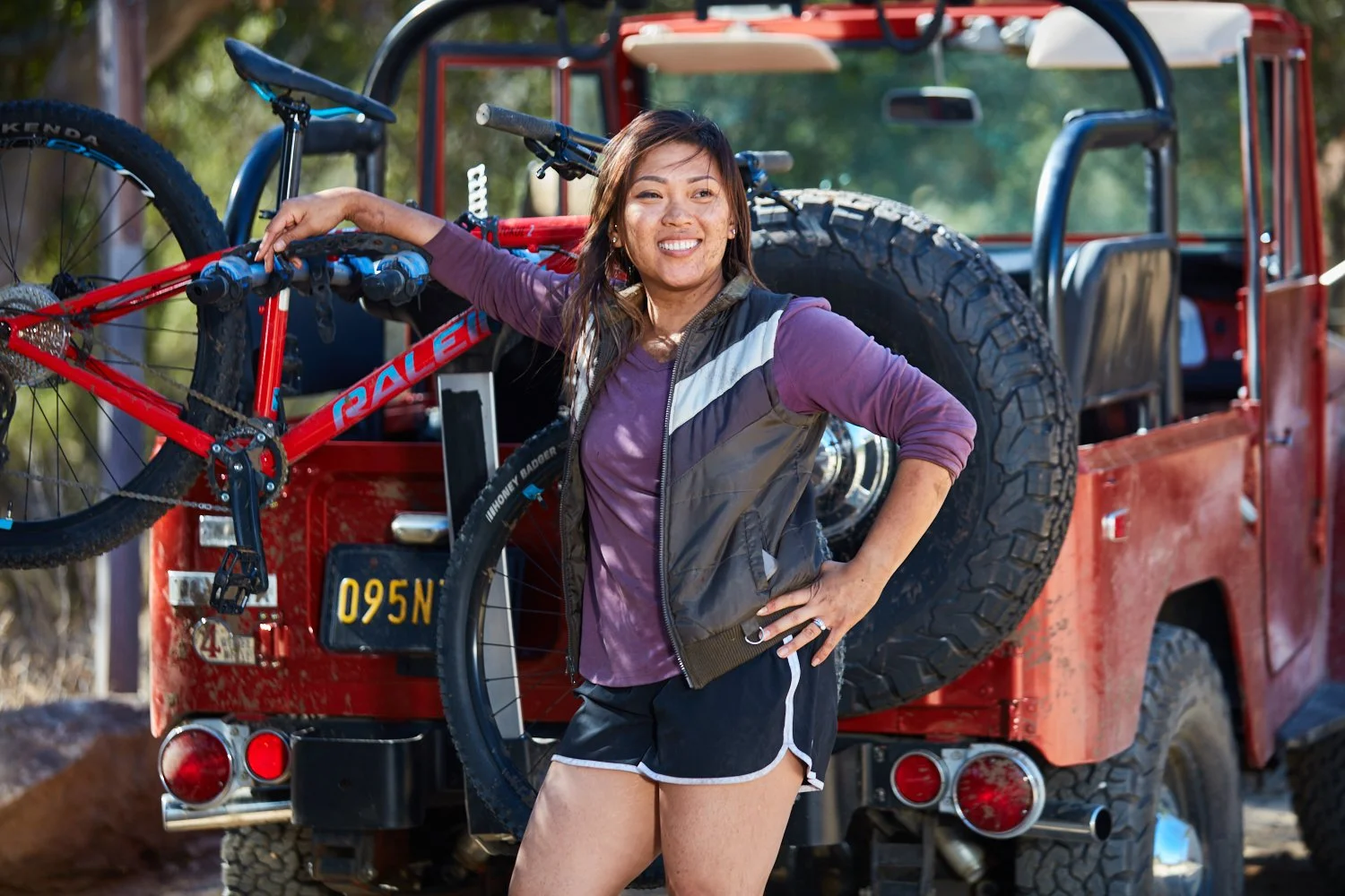 Woman wearing a purple shirt and black shorts with white trim standing next to a red off-road vehicle with a bike mounted on the back, outdoors on a sunny day.