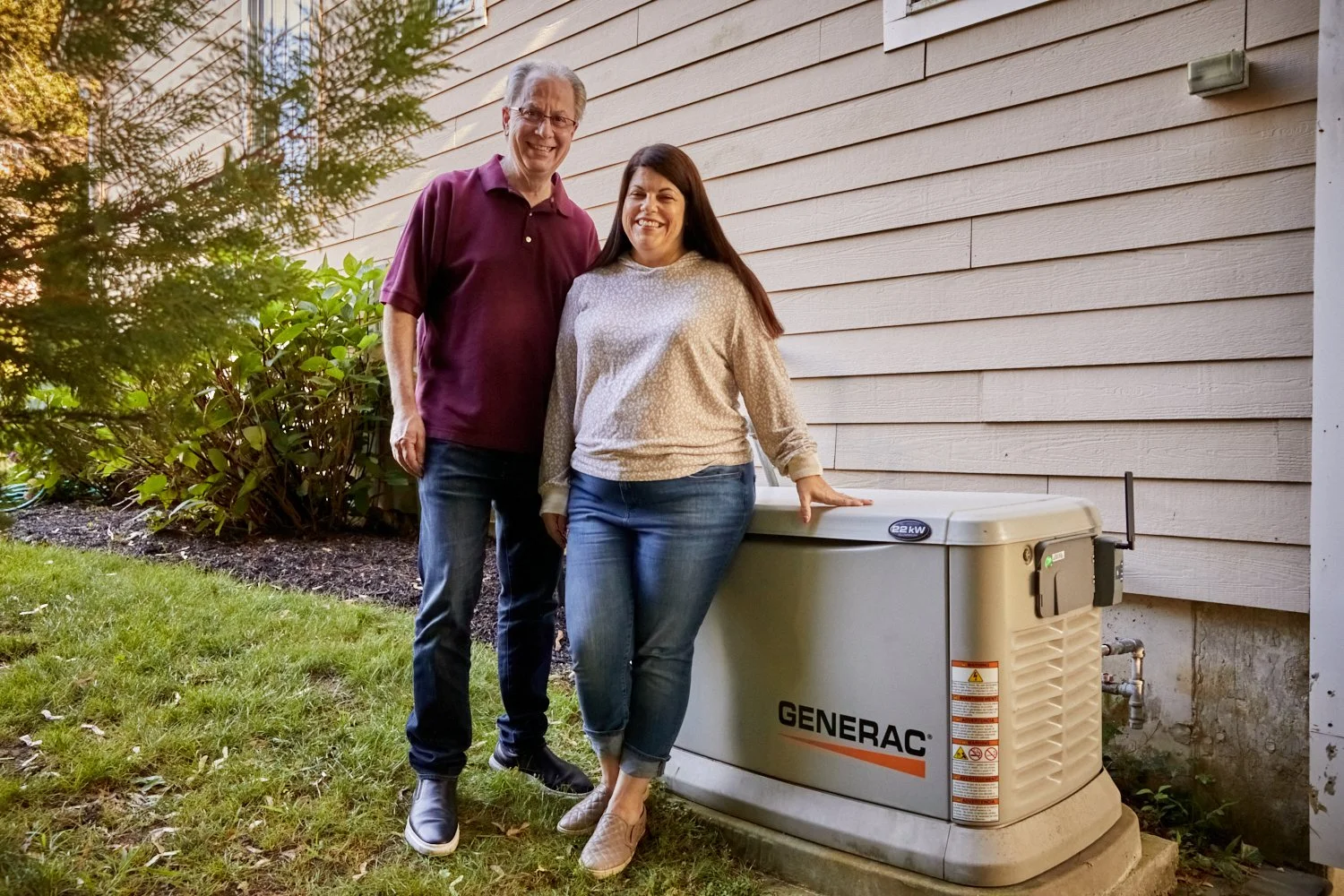 A man and a woman standing next to a Generac generator outside a house on a grassy area. The woman touches the generator with her hand, both are smiling, and there are shrubs and trees in the background.