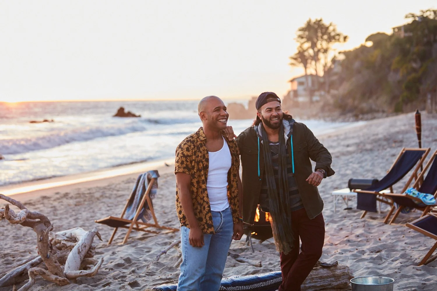 Two men smiling and talking on a beach during sunset, with lounge chairs and trees in the background.