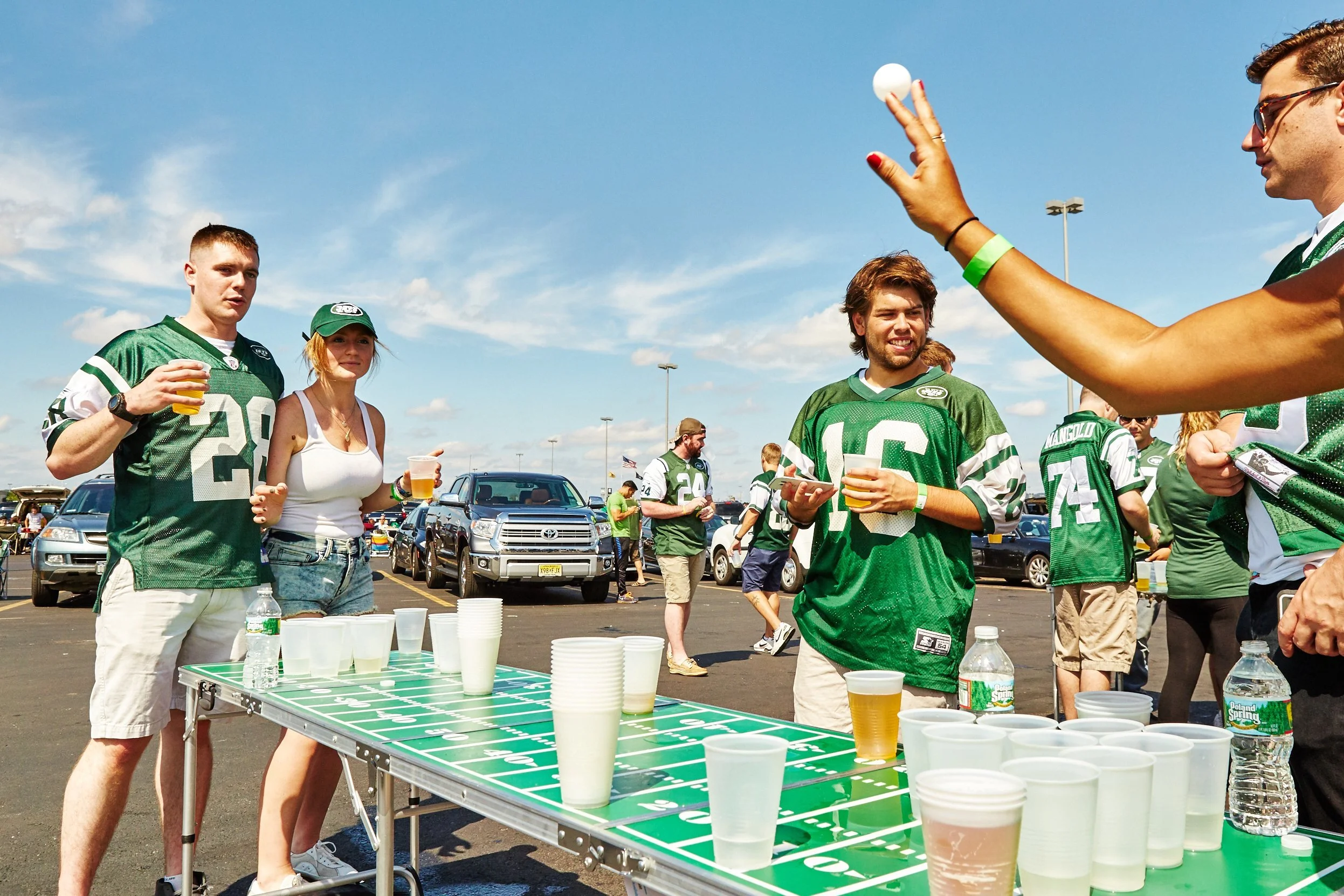 People playing a beer pong game outdoors on a parking lot, wearing green football jerseys, with cars parked behind them and a blue sky overhead.