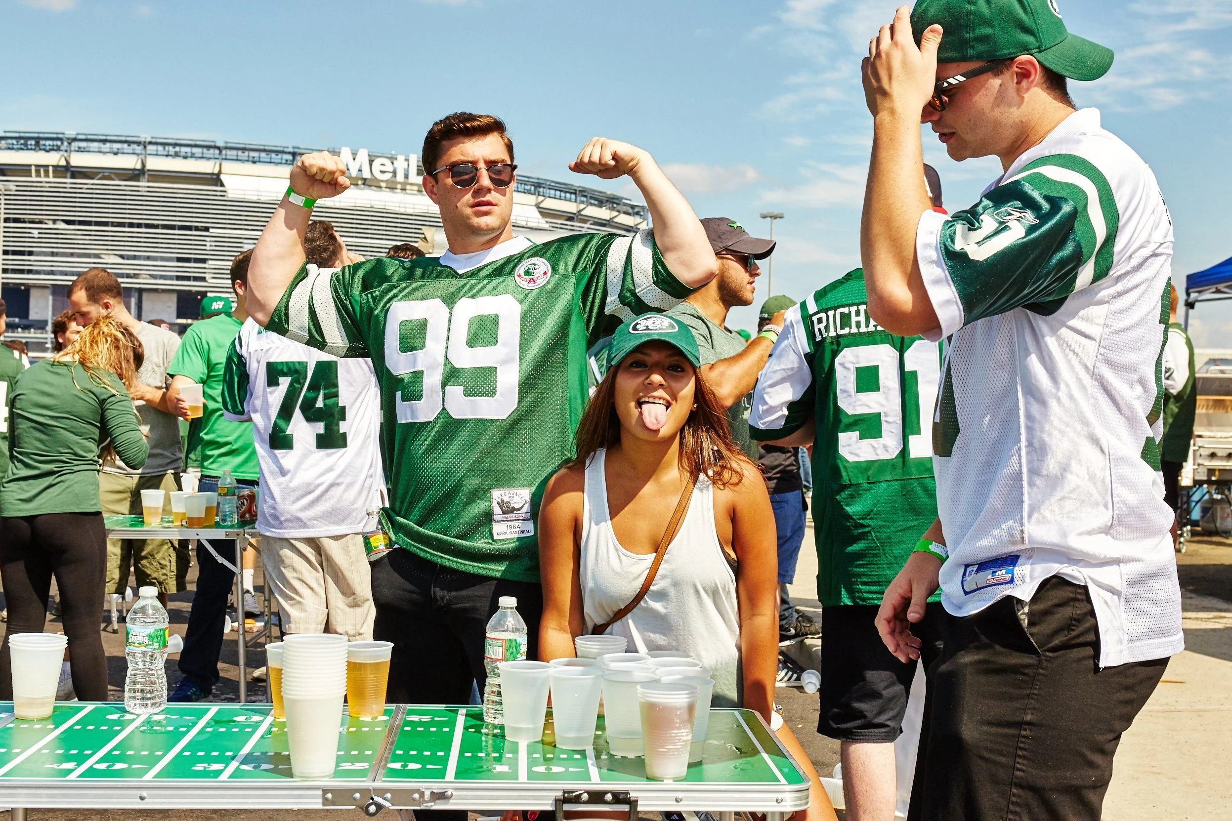 A young woman with long hair sticking out her tongue standing in front of a table with cups filled with beer, surrounded by football fans at a stadium tailgate party.