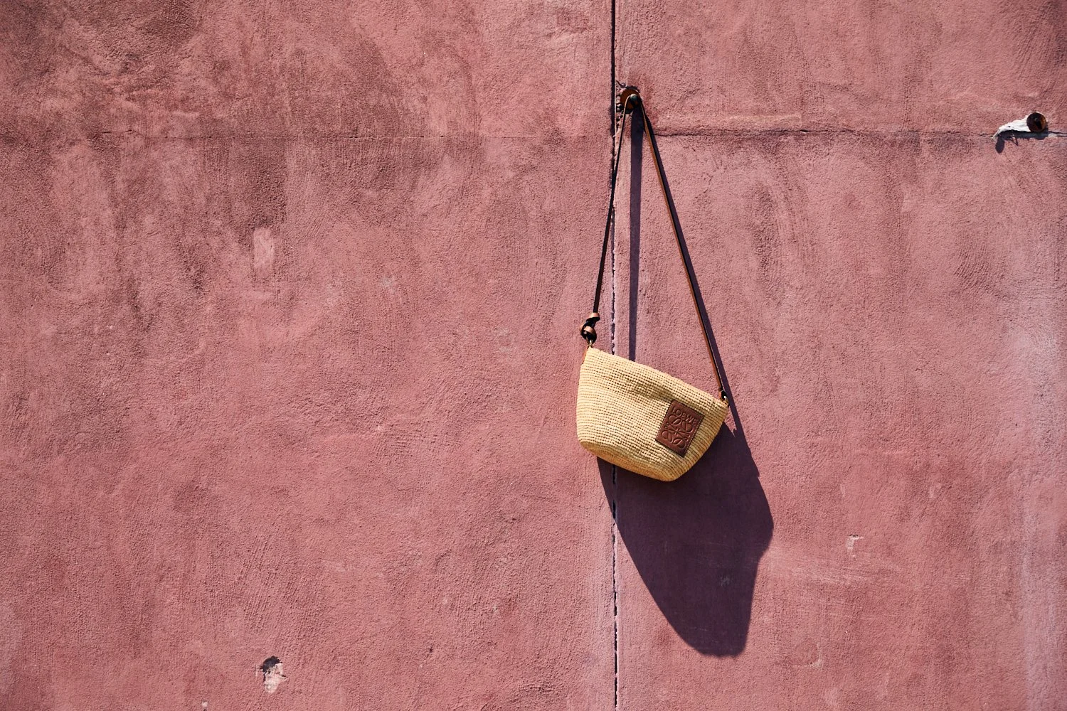 A beige woven bag hanging from a black strap on a pink textured wall, casting a shadow.