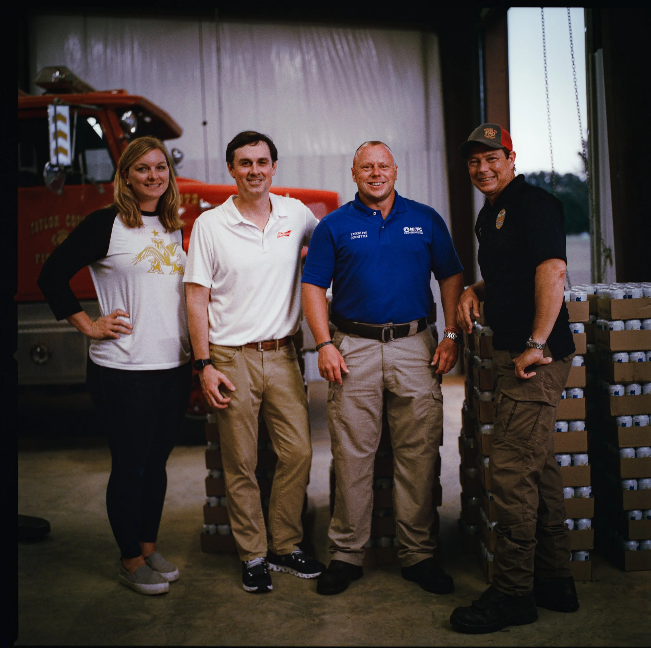 Four people standing in front of a red fire truck inside a garage or workshop, with visible shelves and boxes behind them. The group is smiling and dressed casually.