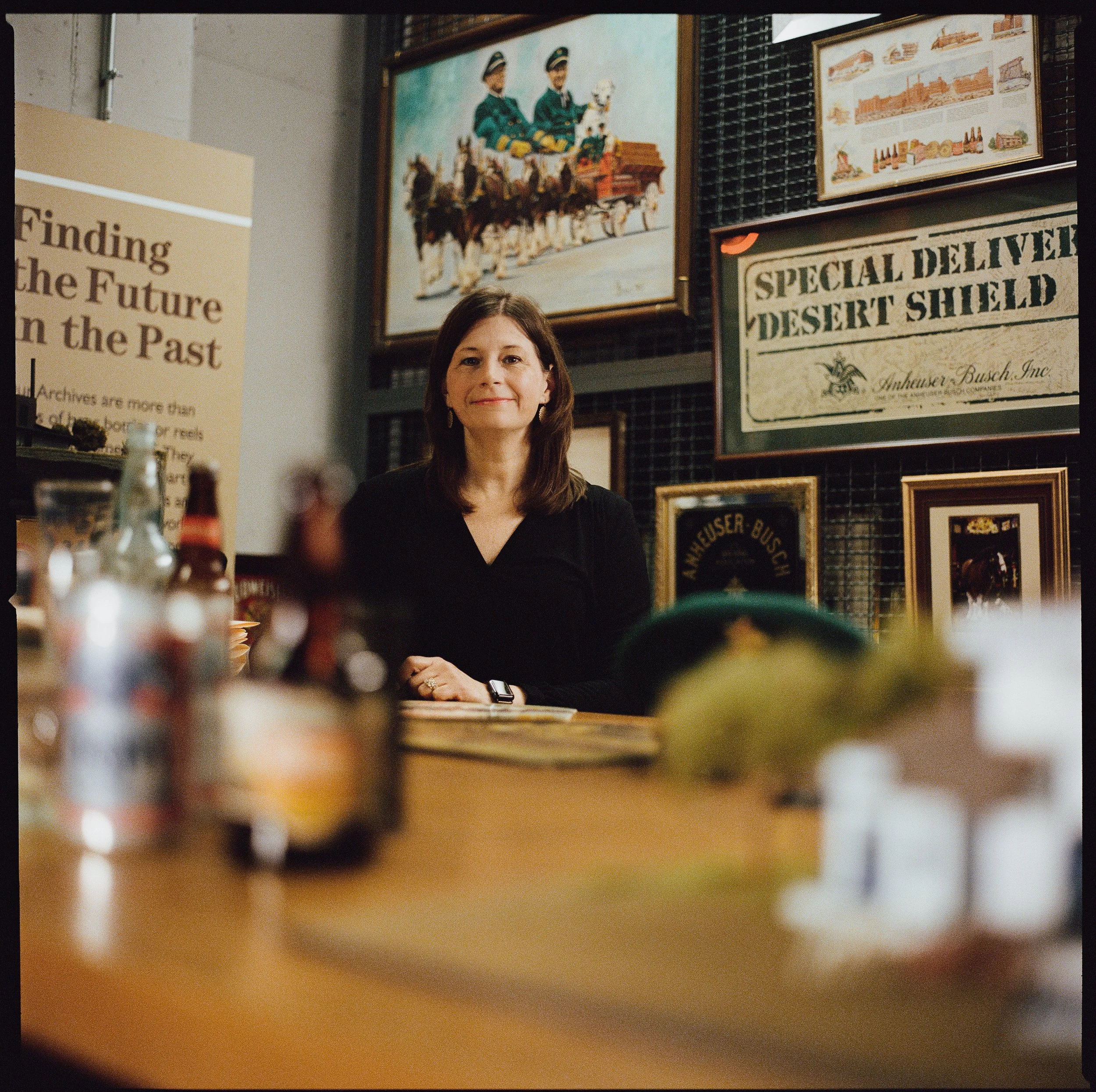 A woman sitting at a table inside a room with framed pictures and signs on the wall behind her. The woman has shoulder-length brown hair and is smiling. The foreground shows blurred objects on the table, including bottles or condiments.