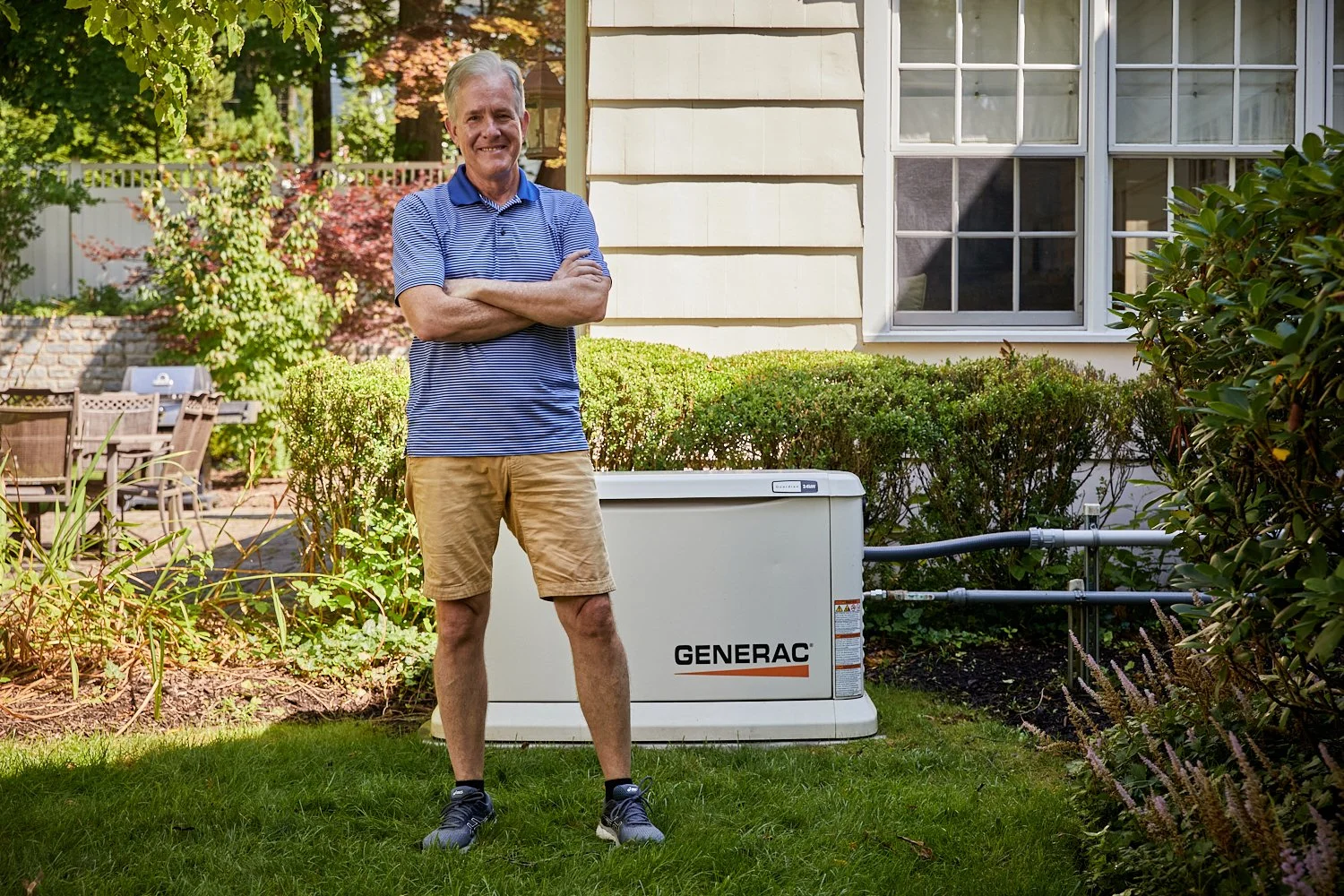 A man standing outdoors on grass with arms crossed, next to a generator labeled 'GENERAC' in a garden area with bushes, trees, and a house with a window in the background.